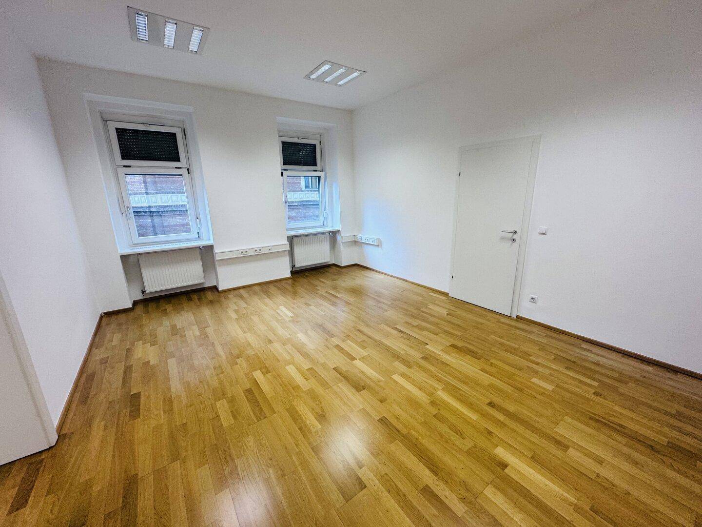 Heller Büroraum mit zwei Fenstern und Holzparkett, ideal für ein kleines Team oder Einzelbüro.