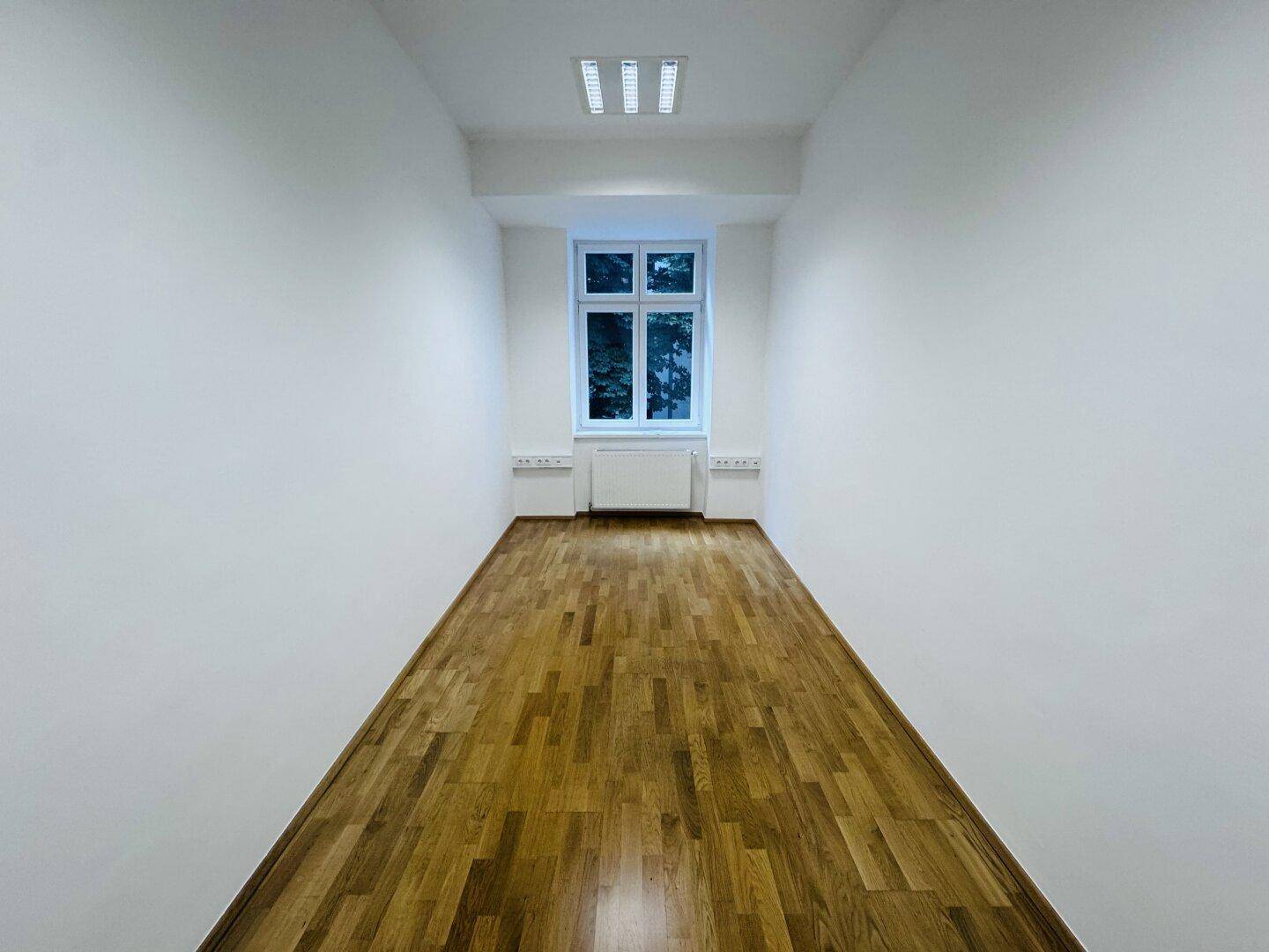 Schmaler Büroraum mit einem Fenster und Holzparkett, ideal für ein Einzelbüro oder Besprechungszimmer.