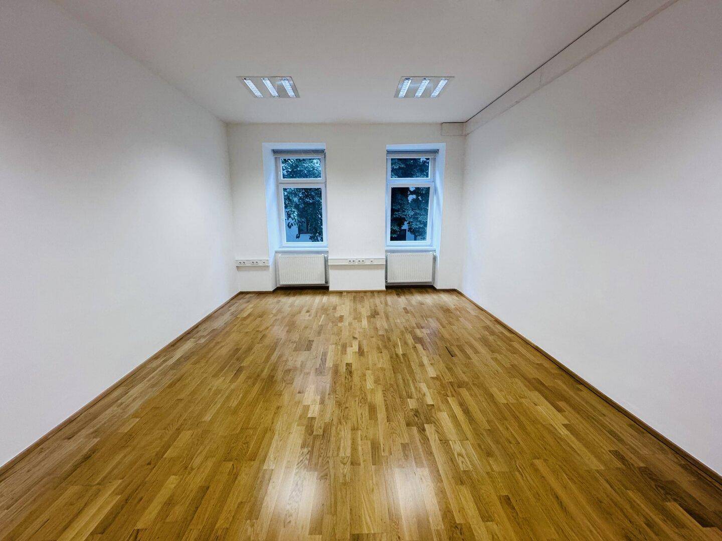 Heller Büroraum mit zwei Fenstern und Holzparkett, bietet viel Tageslicht und eine offene Atmosphäre.