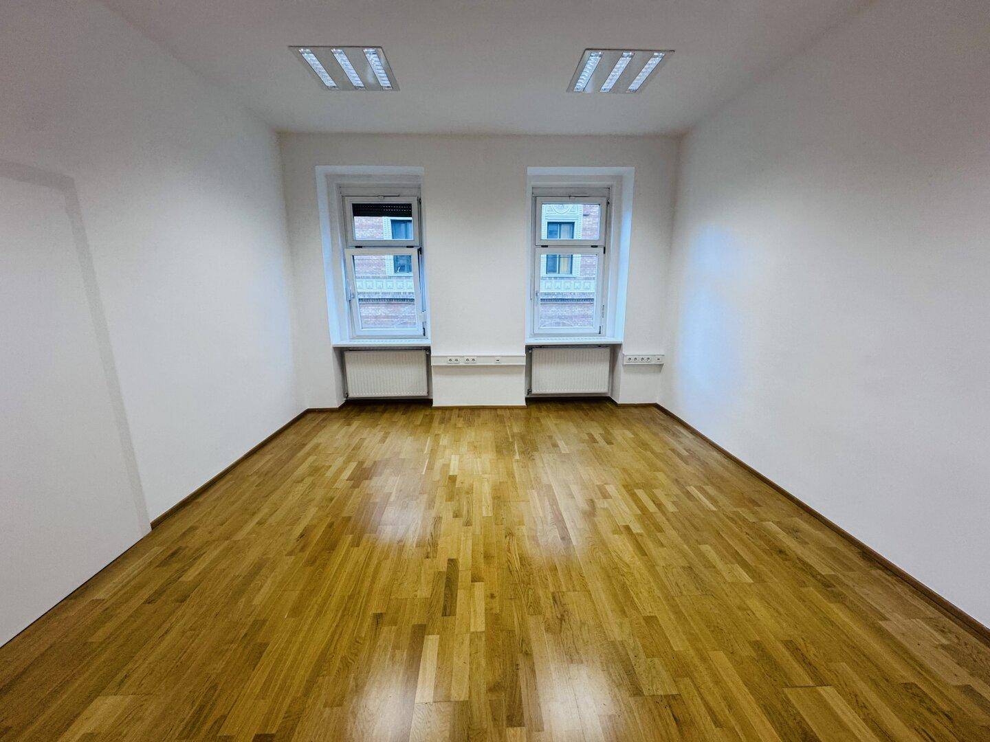 Geräumiger Büroraum mit zwei Fenstern und Holzparkett, ideal für ein Team oder als Großraumbüro.