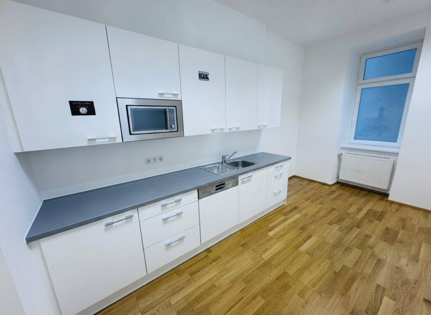 Moderne Einbauküche mit weißen Schränken, grauer Arbeitsplatte und integrierten Geräten, inklusive Mikrowelle und Spülmaschine.
