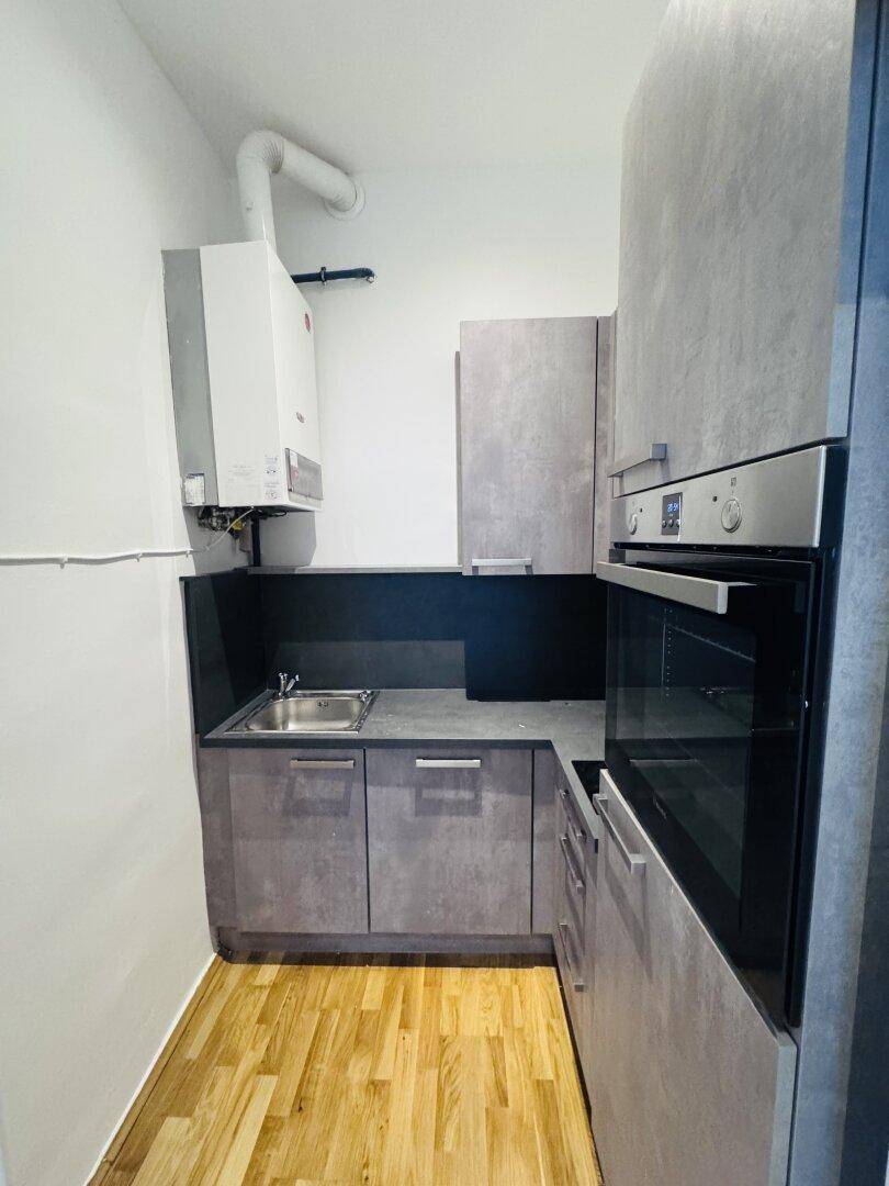 Kleine Küchenzeile mit grauen Schränken, Spüle, Backofen und einem Warmwasserbereiter an der Wand.