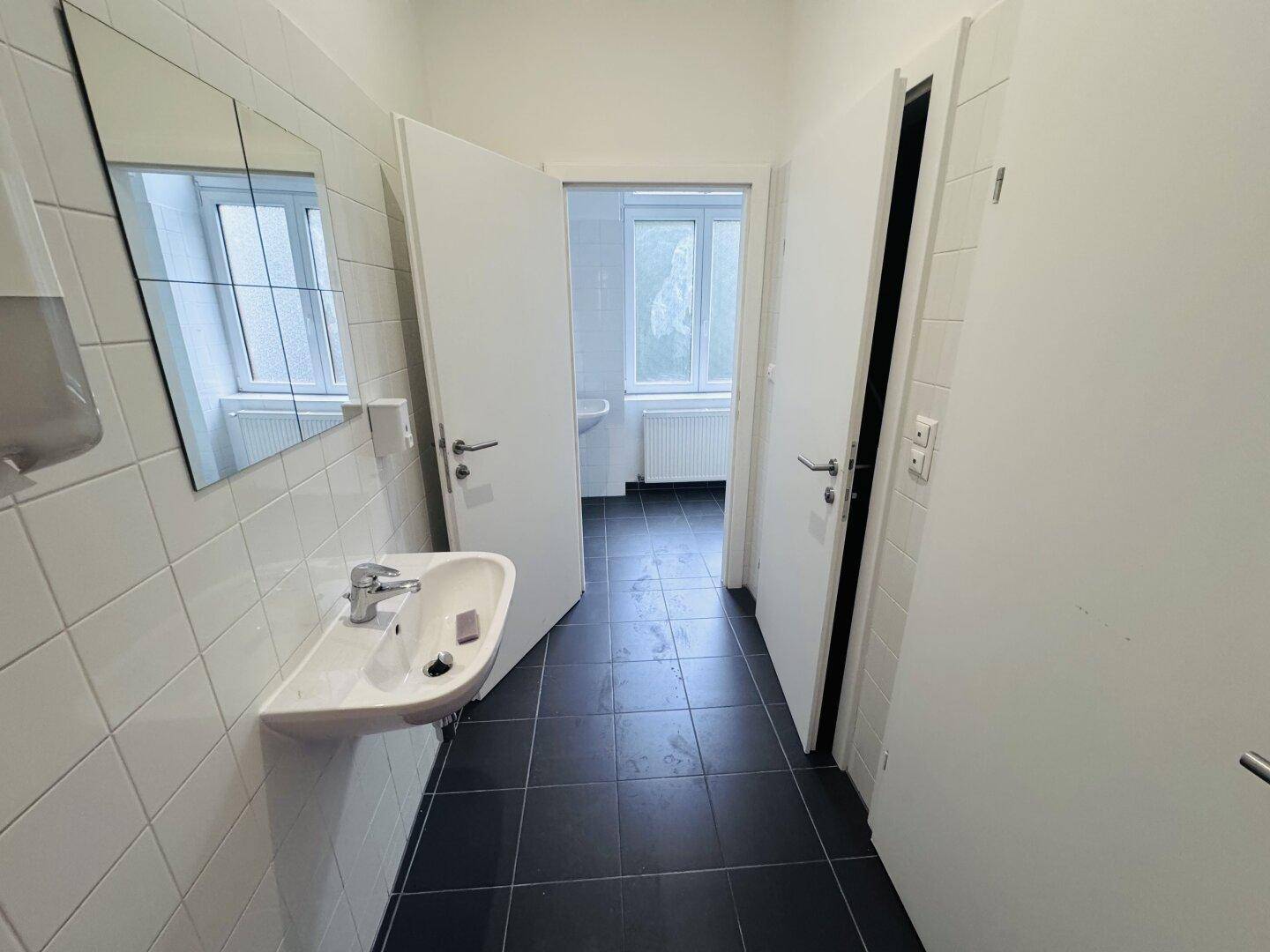 Badezimmer mit dunklen Fliesen, Waschbecken und einem Spiegel, der ein Fenster reflektiert.
