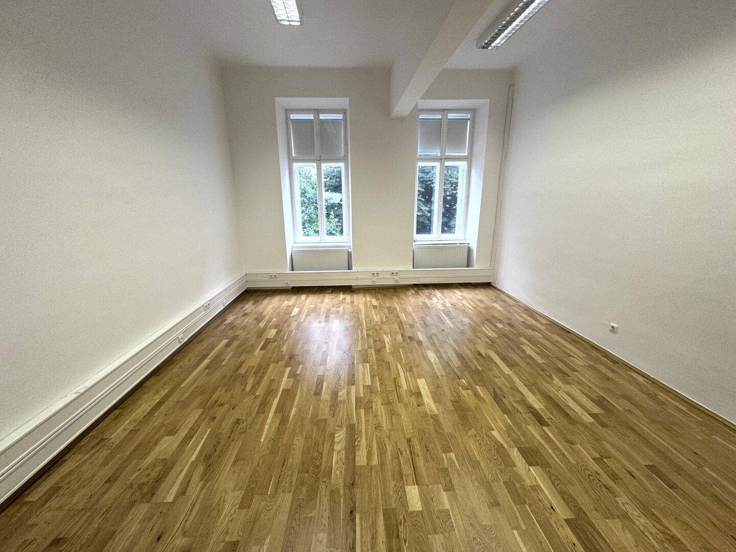 Leerer Büroraum mit Holzboden und zwei hohen Fenstern, die eine grüne Aussicht bieten.