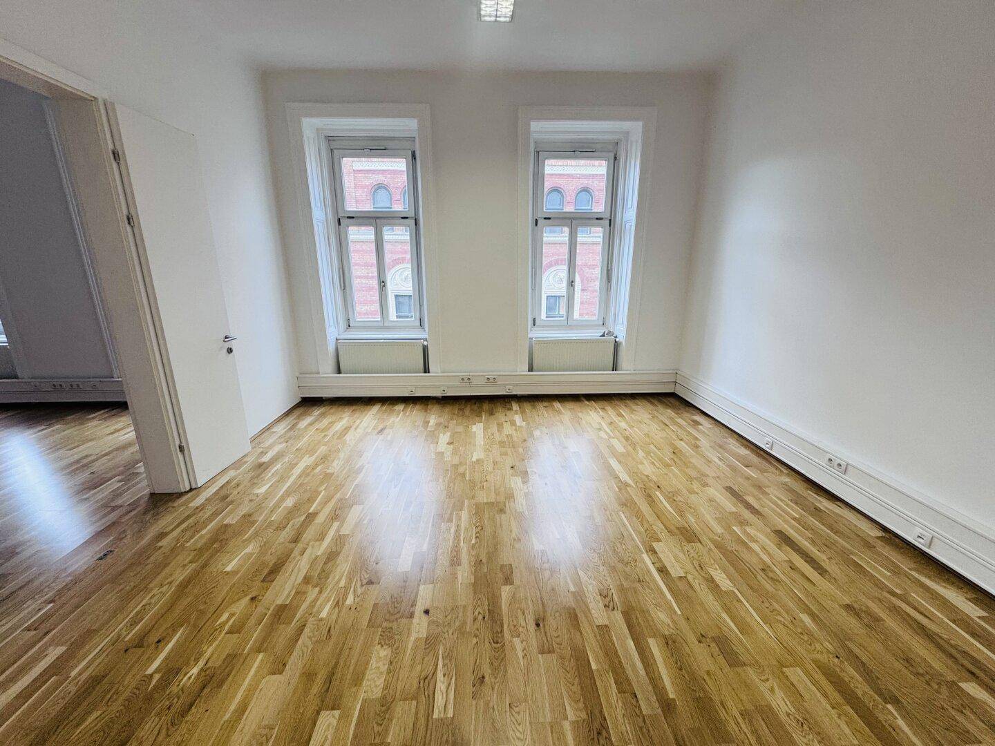 Heller Büroraum mit Holzboden und zwei Fenstern, die einen Blick auf die gegenüberliegenden Gebäude ermöglichen.