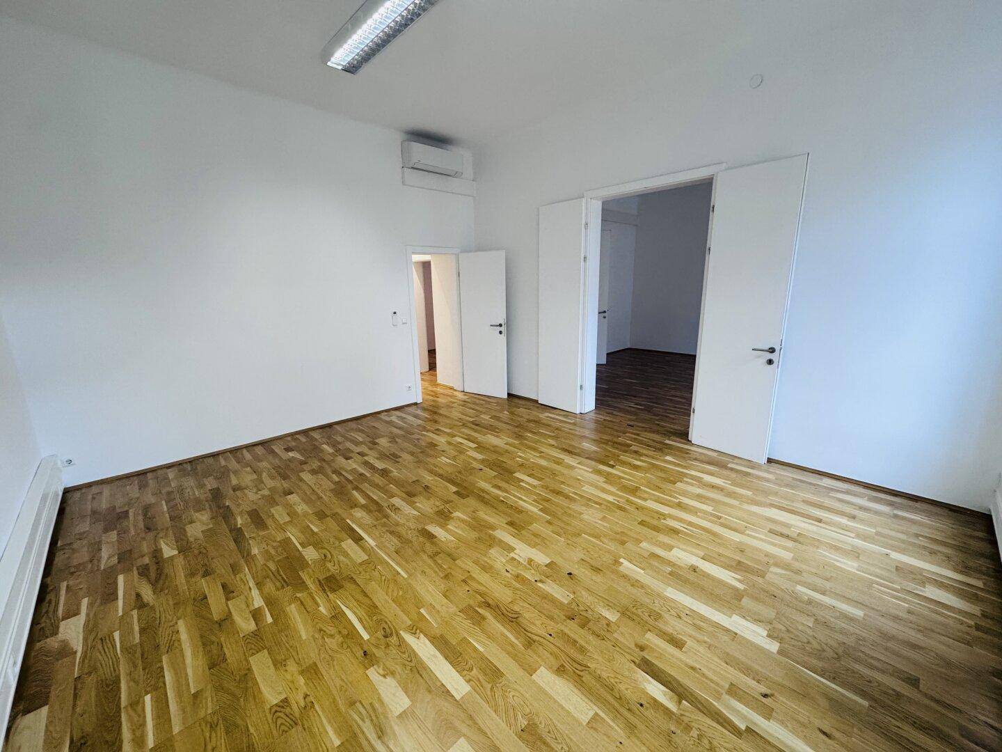 Geräumiger Büroraum mit Holzboden, Klimaanlage und mehreren Türen zu angrenzenden Bereichen.