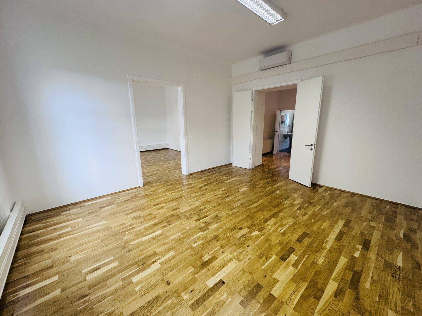 Heller Büroraum mit Holzboden, Klimaanlage und Zugang zu weiteren Räumen.