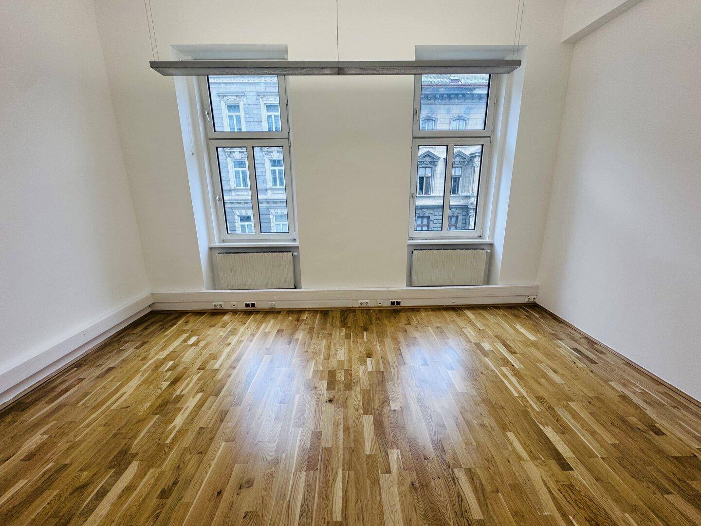Heller Büroraum mit Holzboden und zwei Fenstern, die einen Blick auf die Stadt bieten.