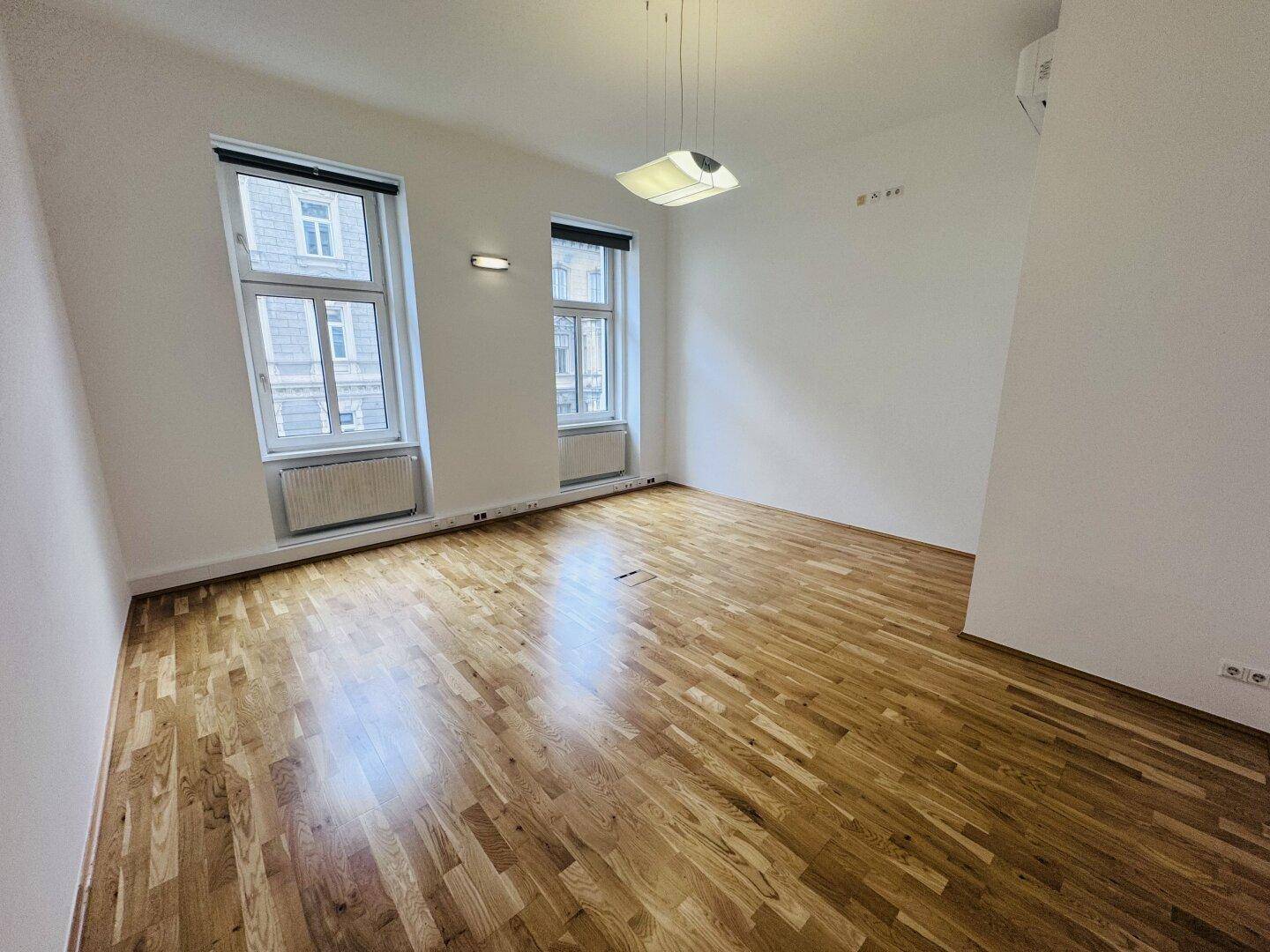 Büroraum mit Holzboden, Klimaanlage und zwei Fenstern, die viel Tageslicht hereinlassen.