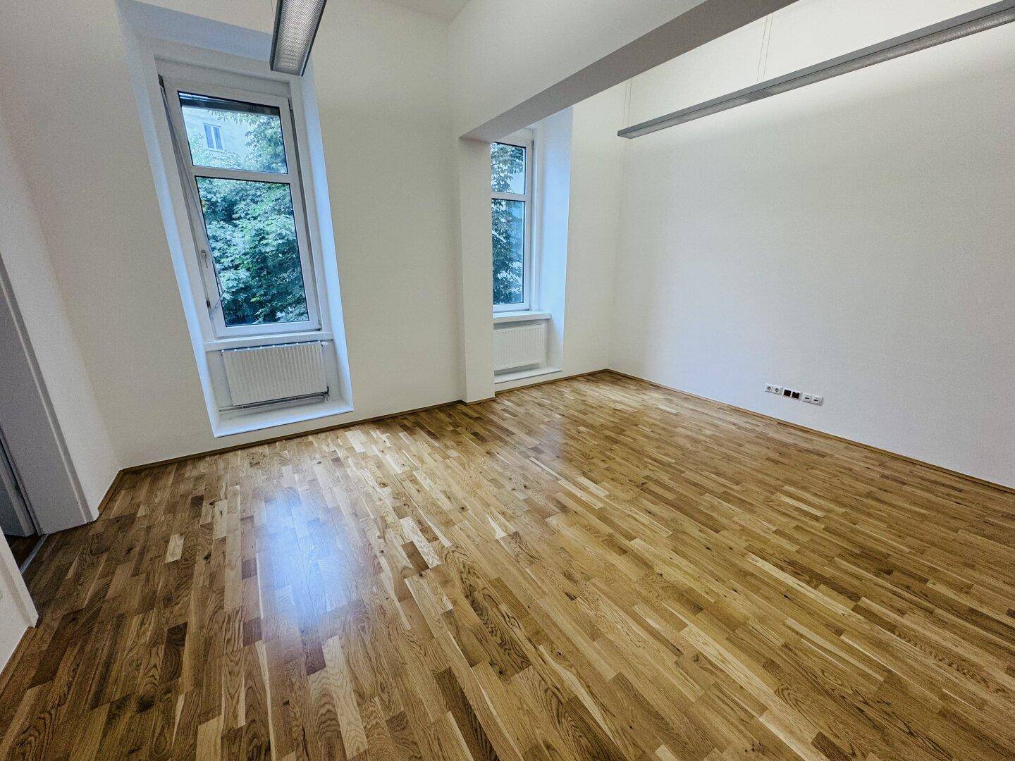 Heller Büroraum mit Holzboden und zwei Fenstern, die eine grüne Aussicht bieten.