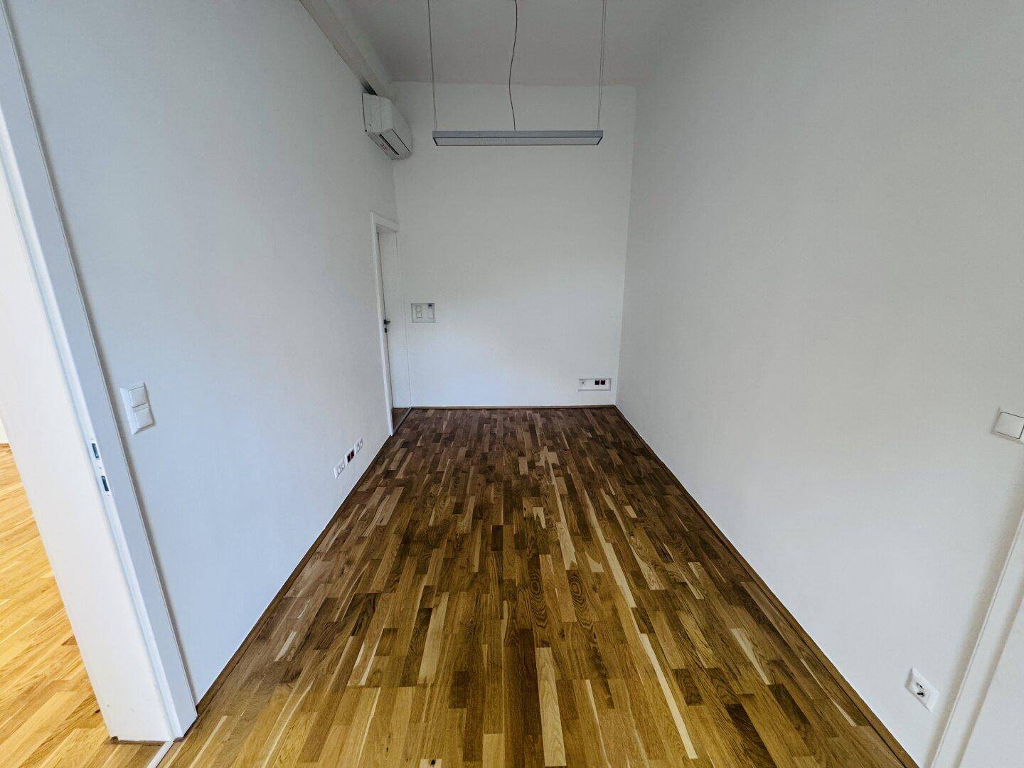Schmaler Büroraum mit Holzboden und einer Klimaanlage an der Wand.