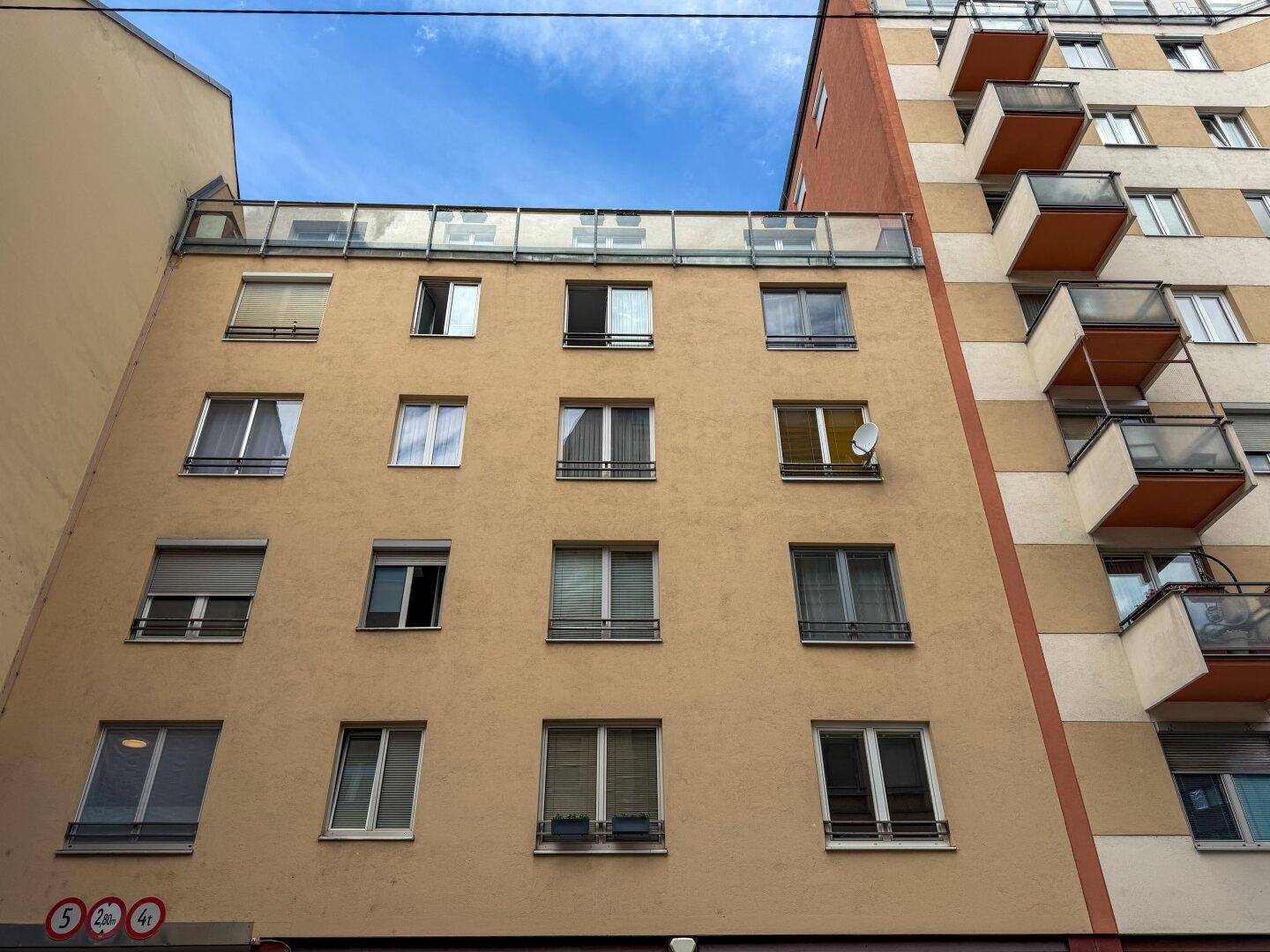 Fassade eines Wohnhauses mit zahlreichen Fenstern und Rollläden, angrenzend an ein Gebäude mit Balkonen.
