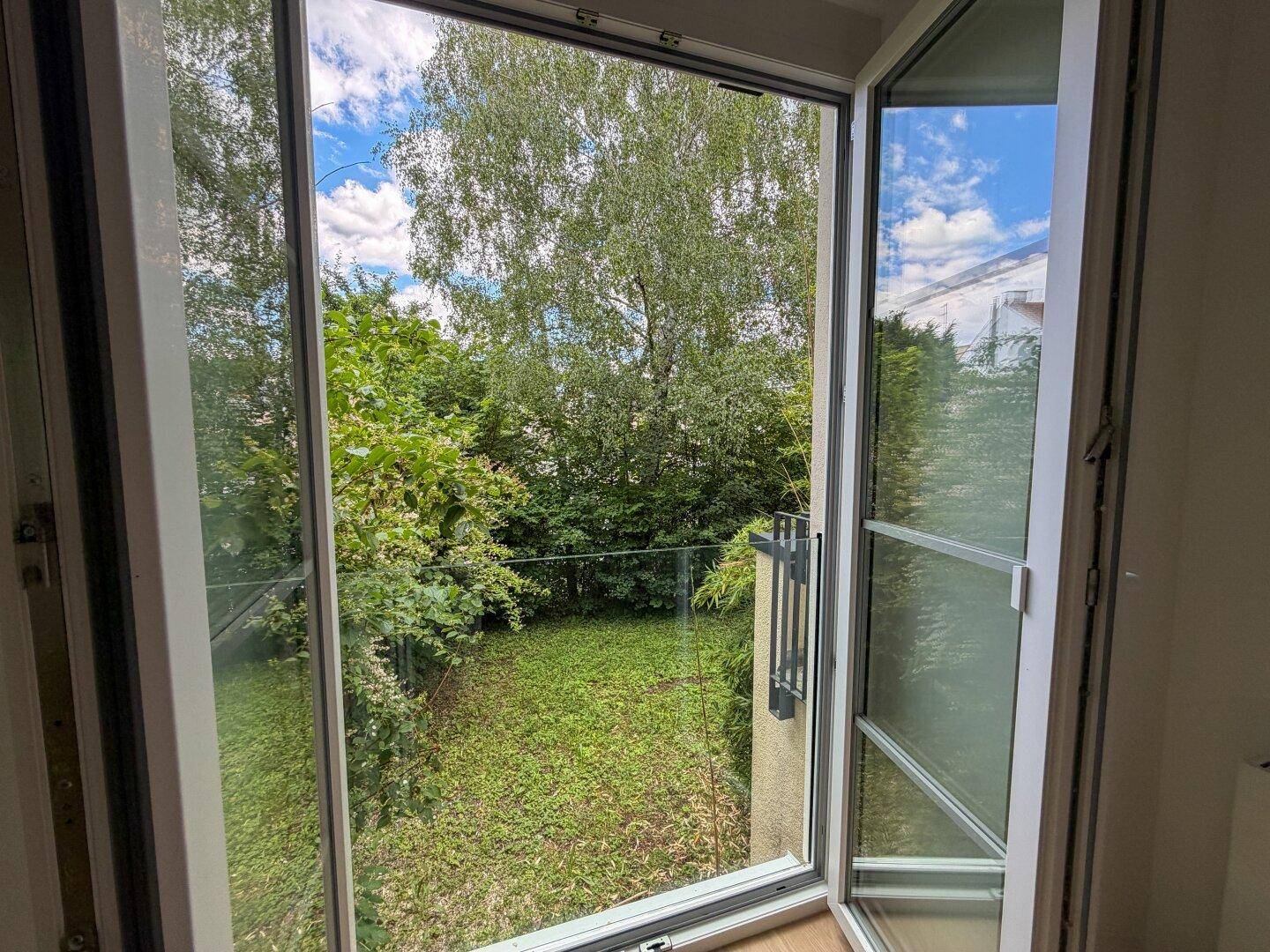 Blick durch die Balkontür auf einen kleinen, grünen Gartenbereich mit Bäumen und blauem Himmel.