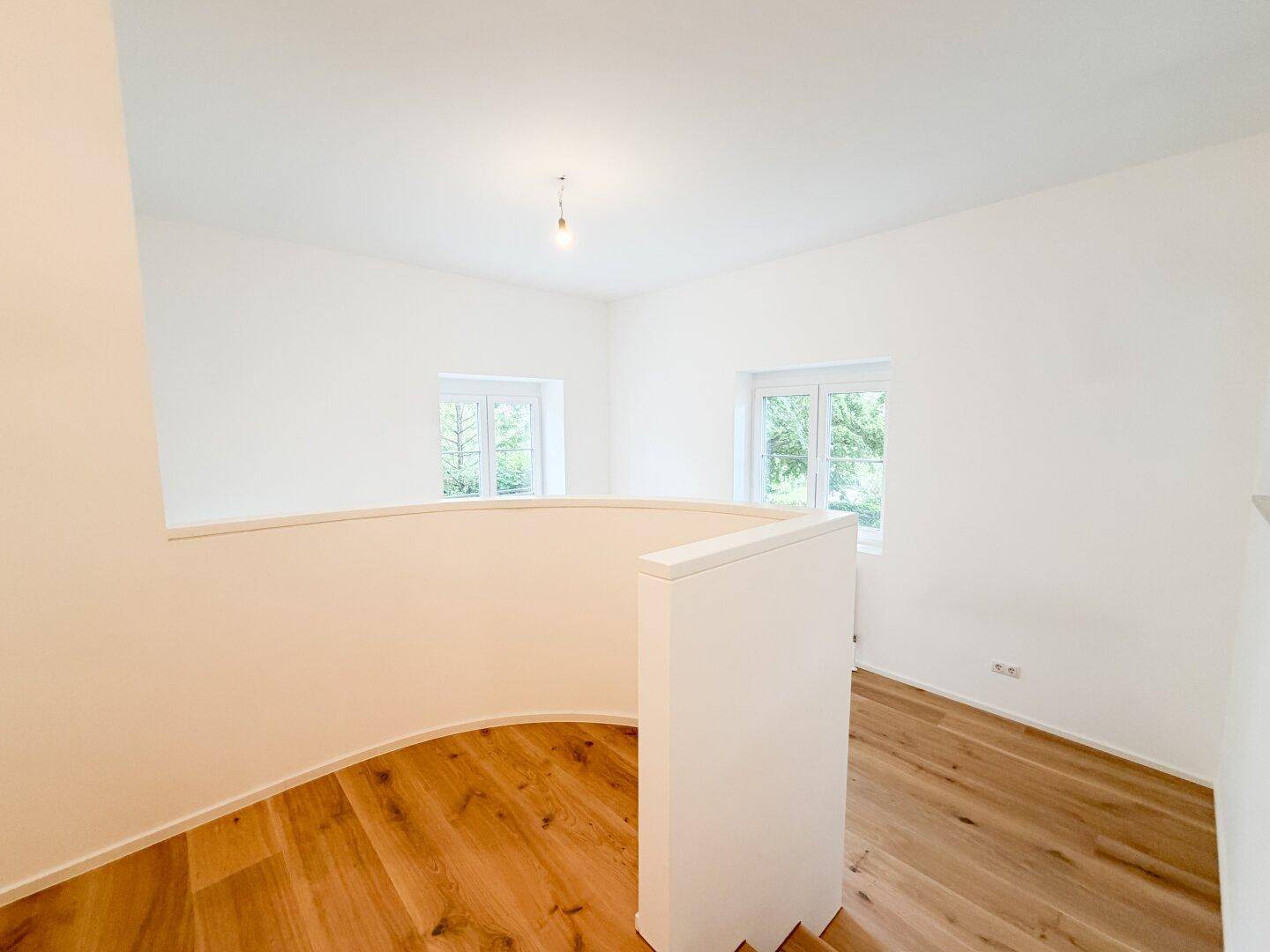 Heller Flurbereich mit geschwungener Wand, Holzfußboden und zwei Fenstern mit Blick ins Grüne.
