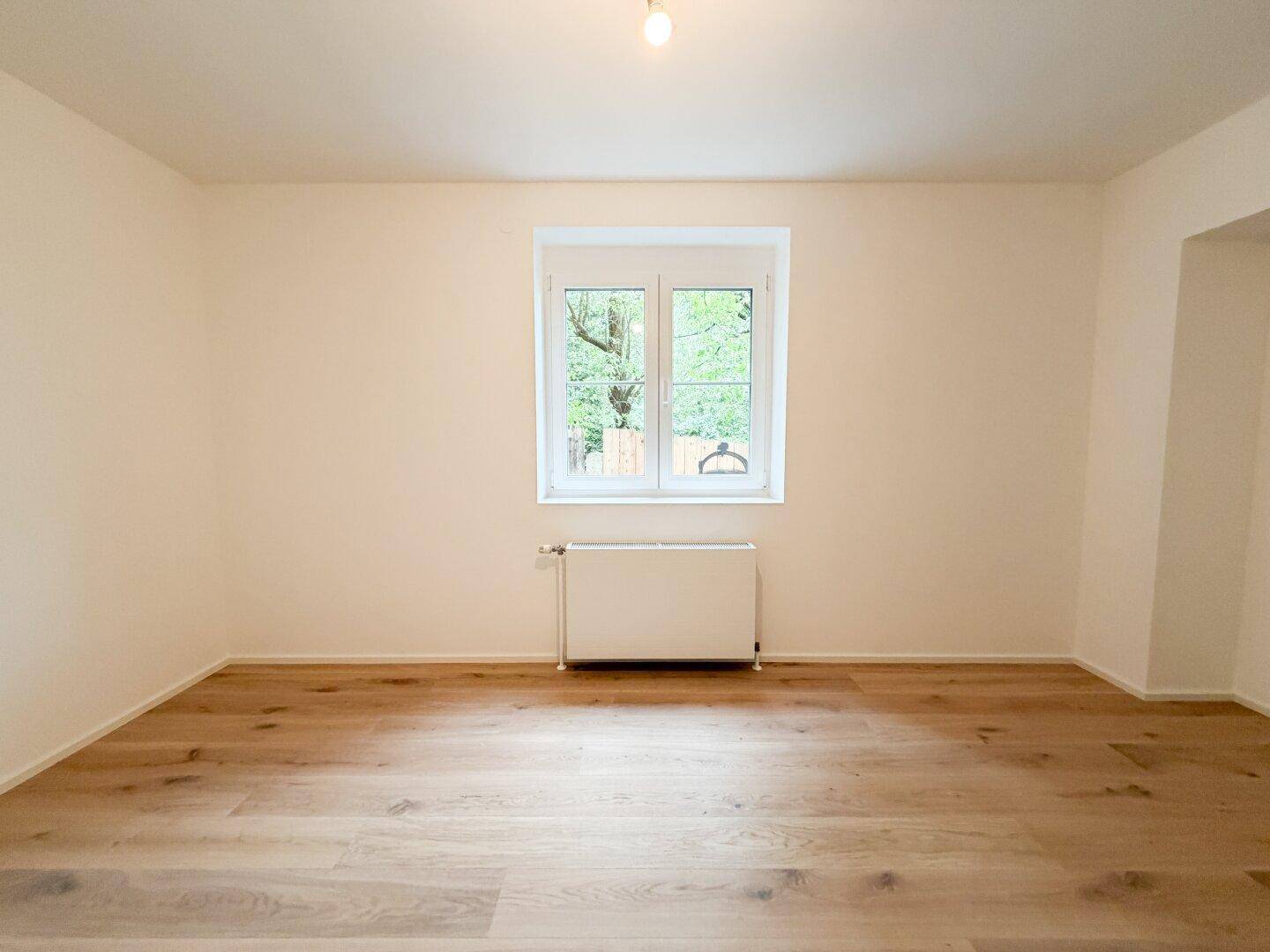Geräumiger, heller Raum mit neuem Holzfußboden und Blick ins Grüne durch das Fenster.