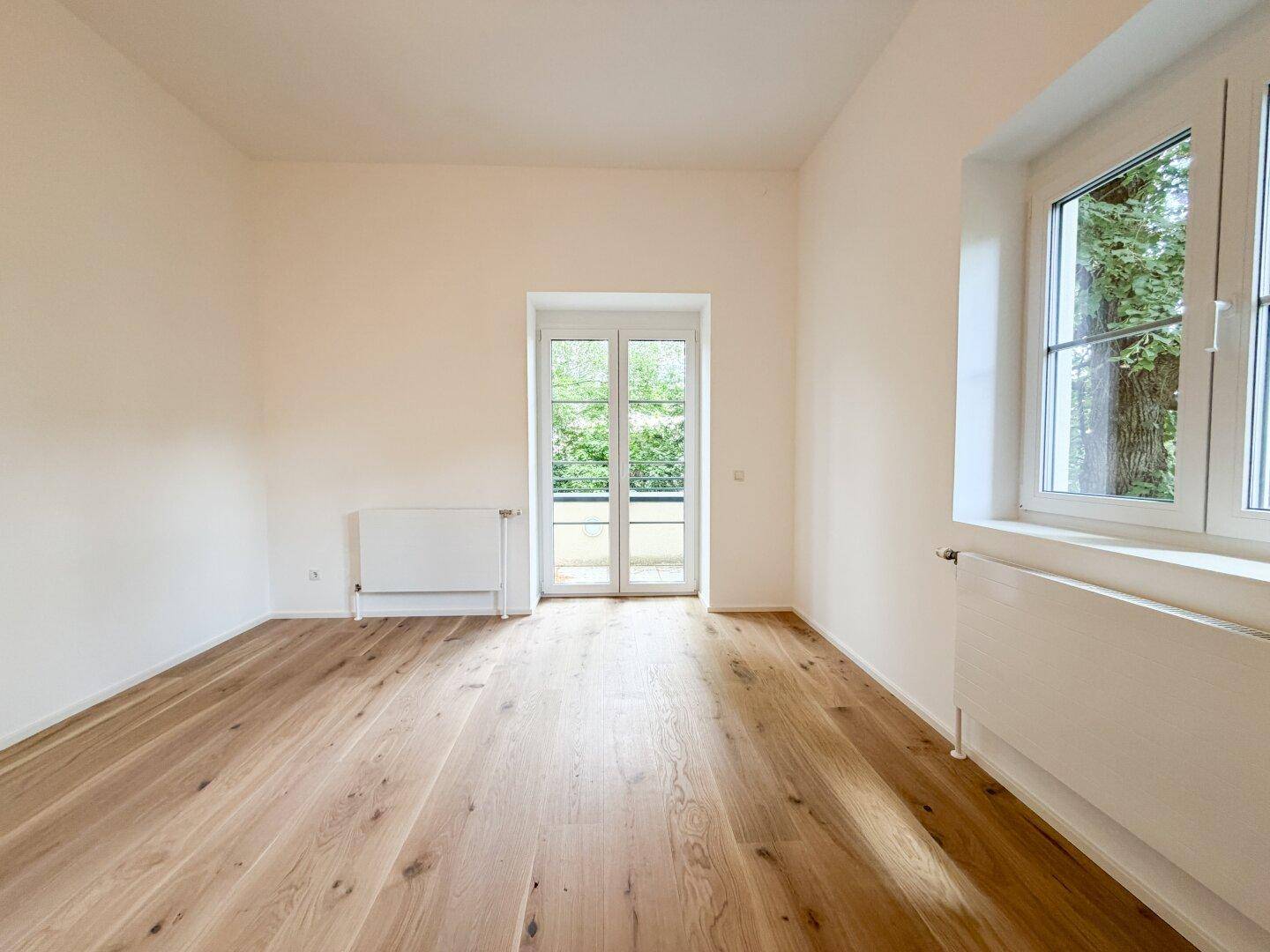 Heller Raum mit Holzfußboden, Fenster und Zugang zu einem französischen Balkon.