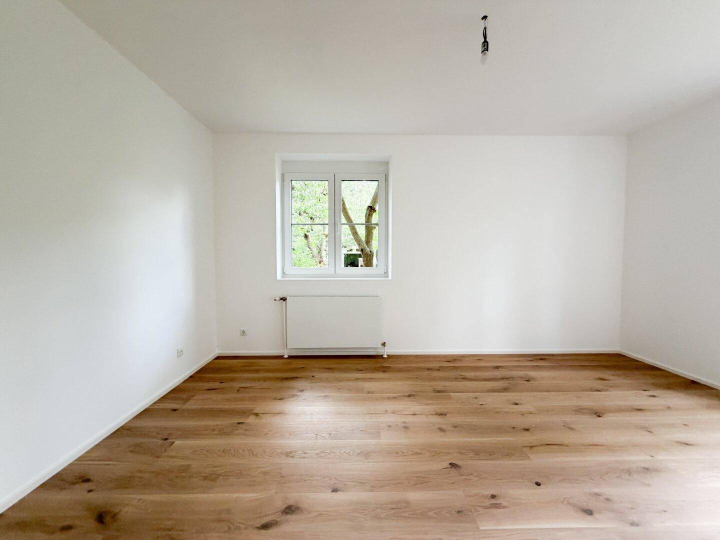 Geräumiger, heller Raum mit neuem Holzfußboden und einem Fenster mit Blick ins Grüne.