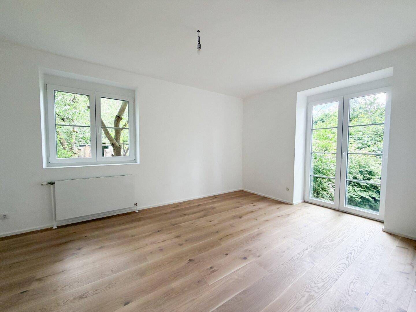 Heller Raum mit Holzfußboden, Fenster und französischen Balkontüren, die viel Licht hereinlassen.