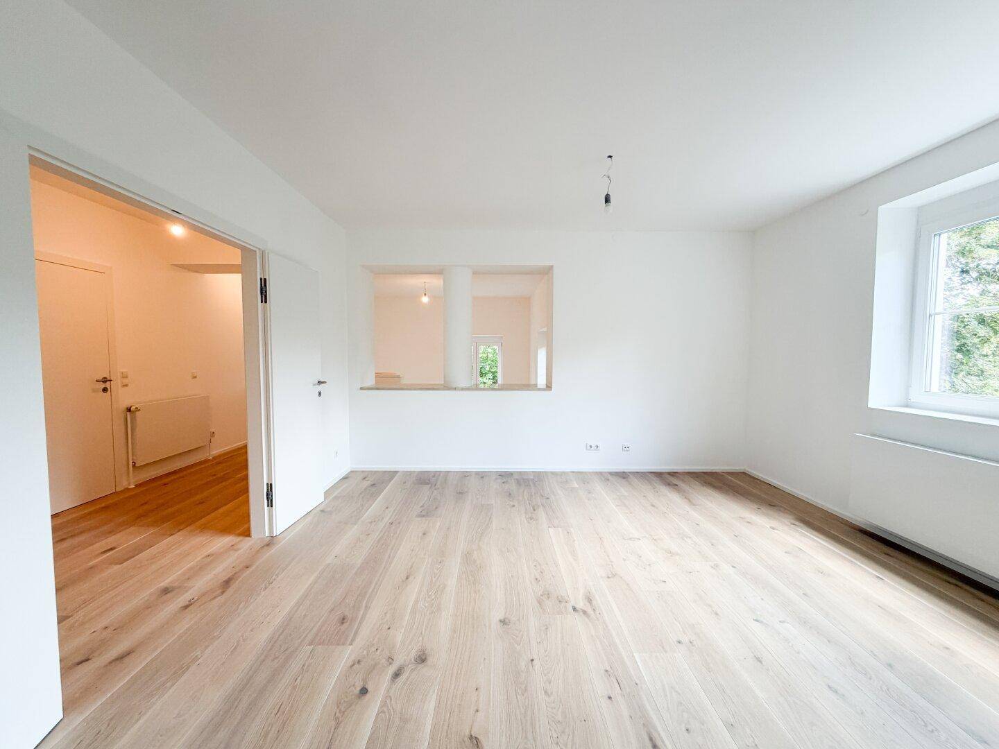 Heller Raum mit Holzfußboden, weißen Wänden und einer offenen Verbindung zu einem weiteren Bereich.