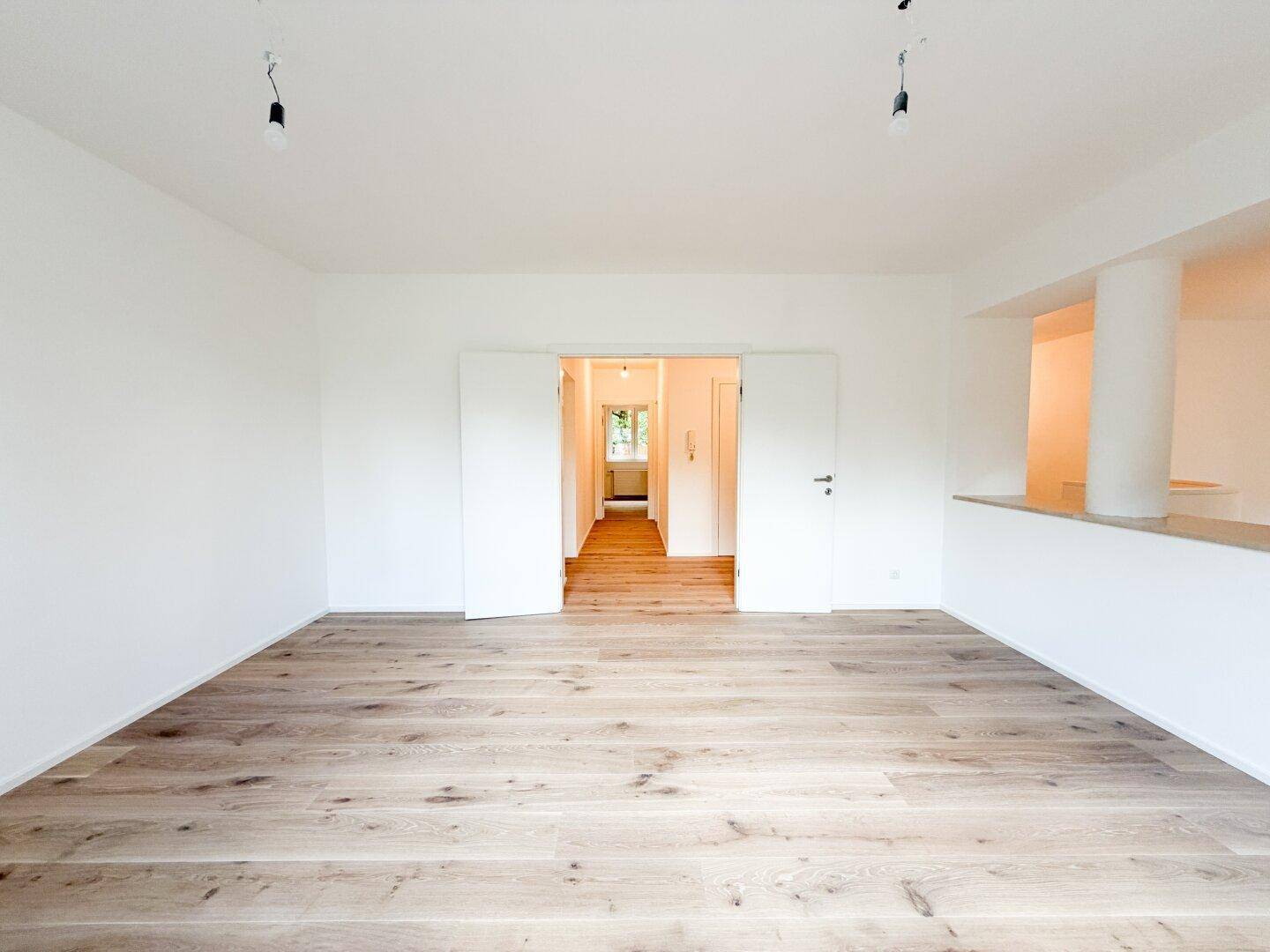 Großer, heller Raum mit Holzfußboden und Doppeltüren, die zu einem Flur führen.