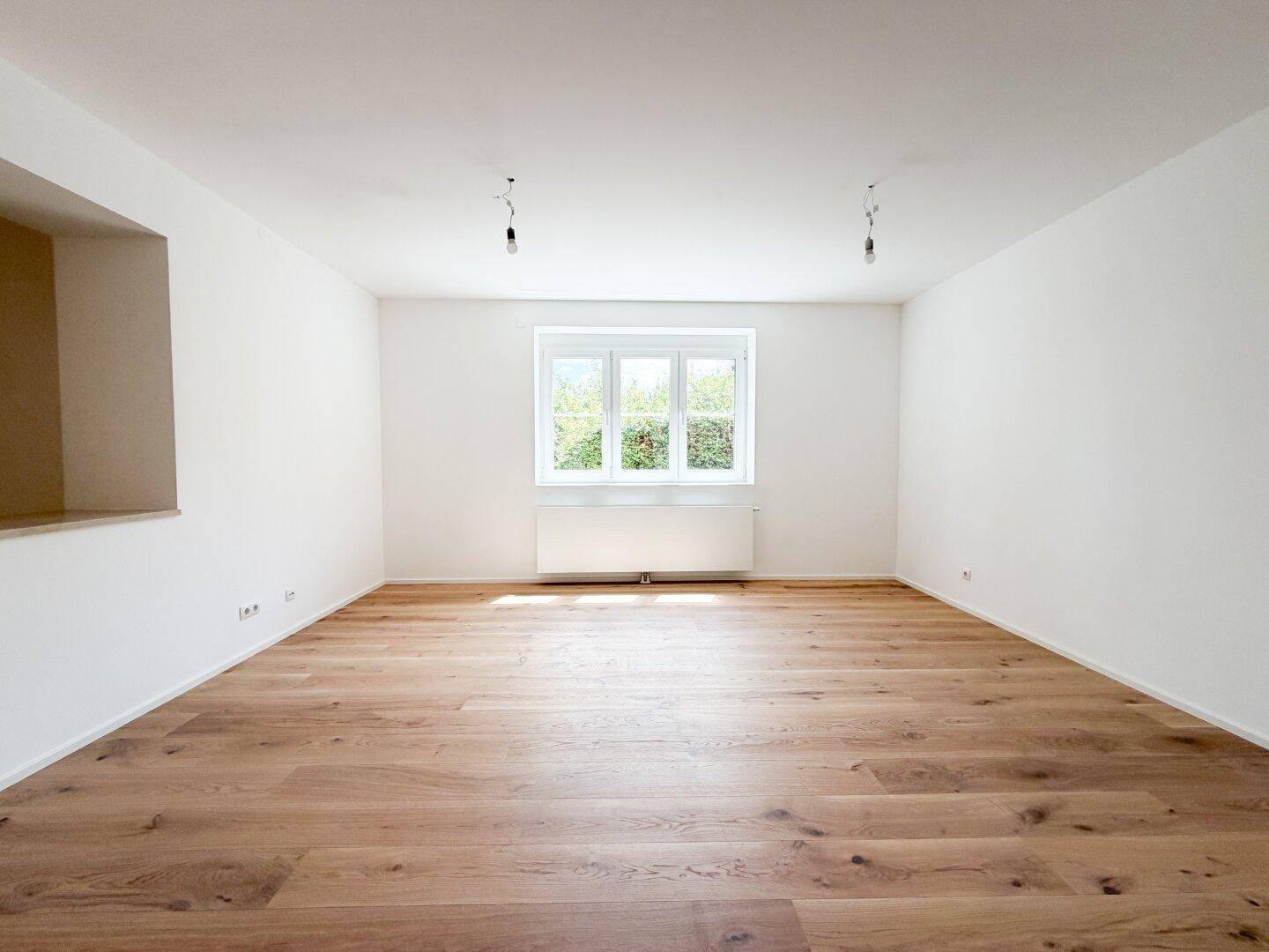 Heller und leerer Raum mit schönem Holzparkett und einem großen Fenster mit Blick ins Grüne.