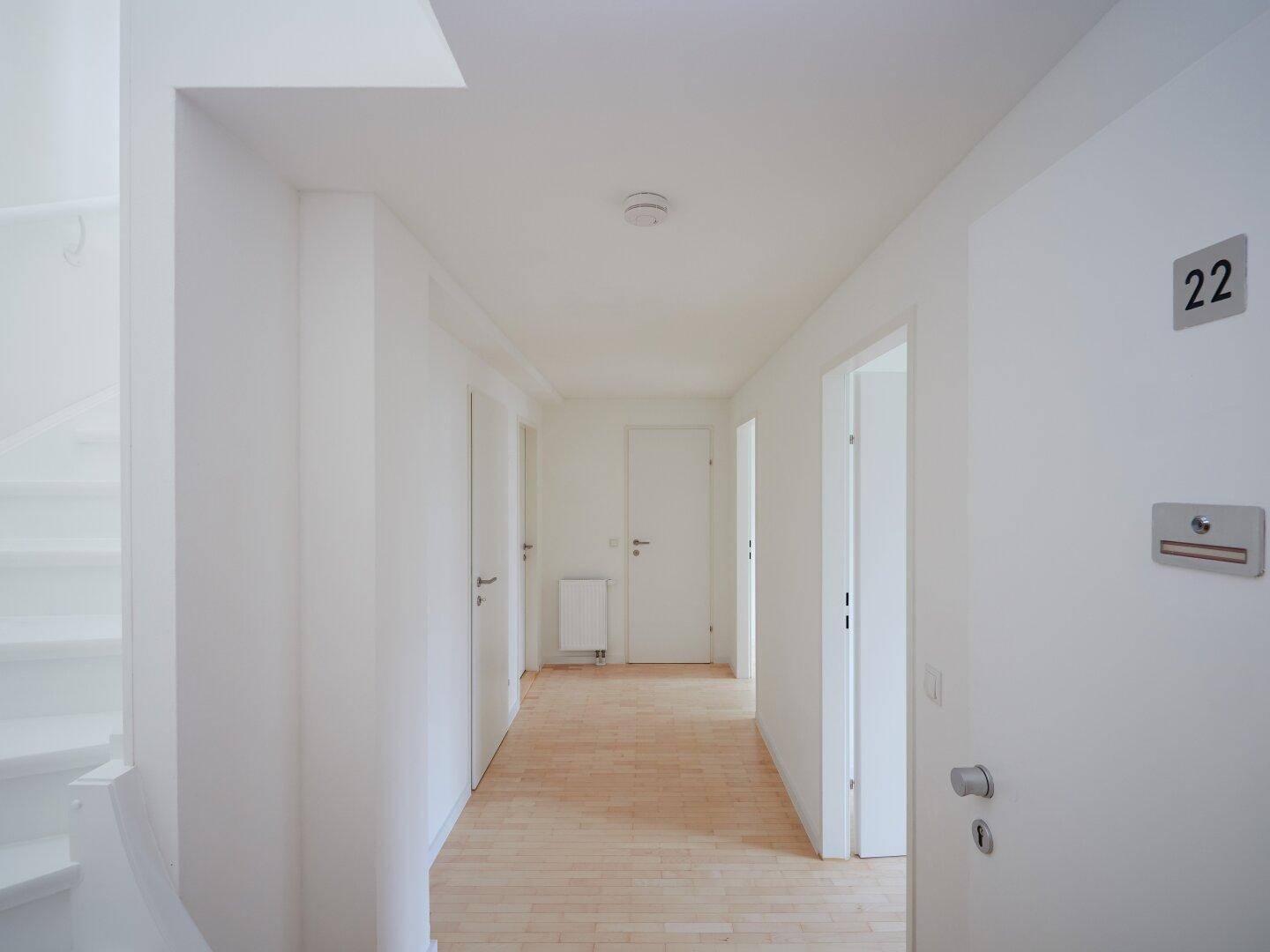 Heller Flur mit Holzboden, weißen Wänden und Zugang zu mehreren Zimmern sowie einem Treppenhaus.