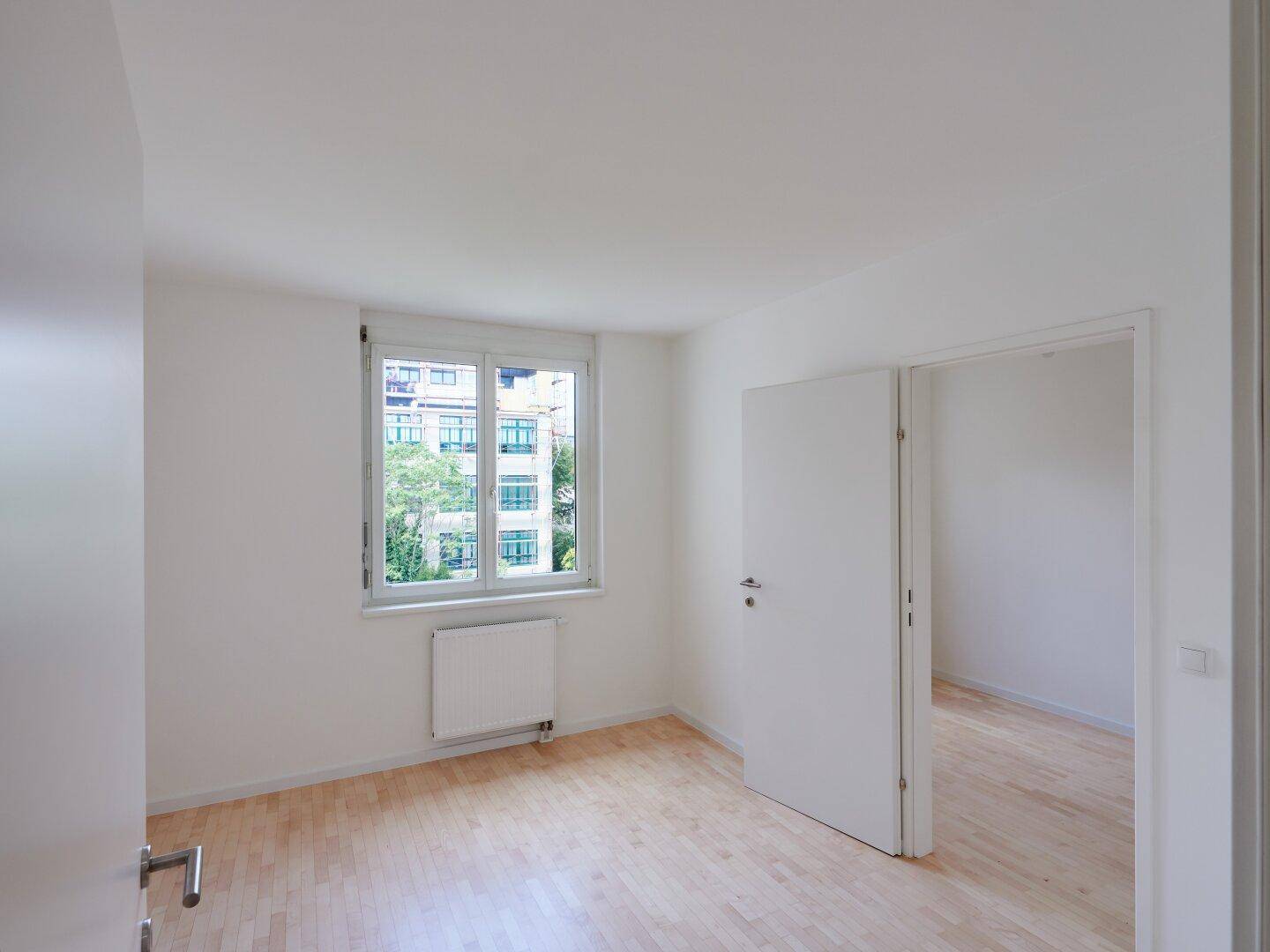 Leeres Schlafzimmer mit hellem Holzboden, weißen Wänden und einem Fenster mit Heizkörper darunter.