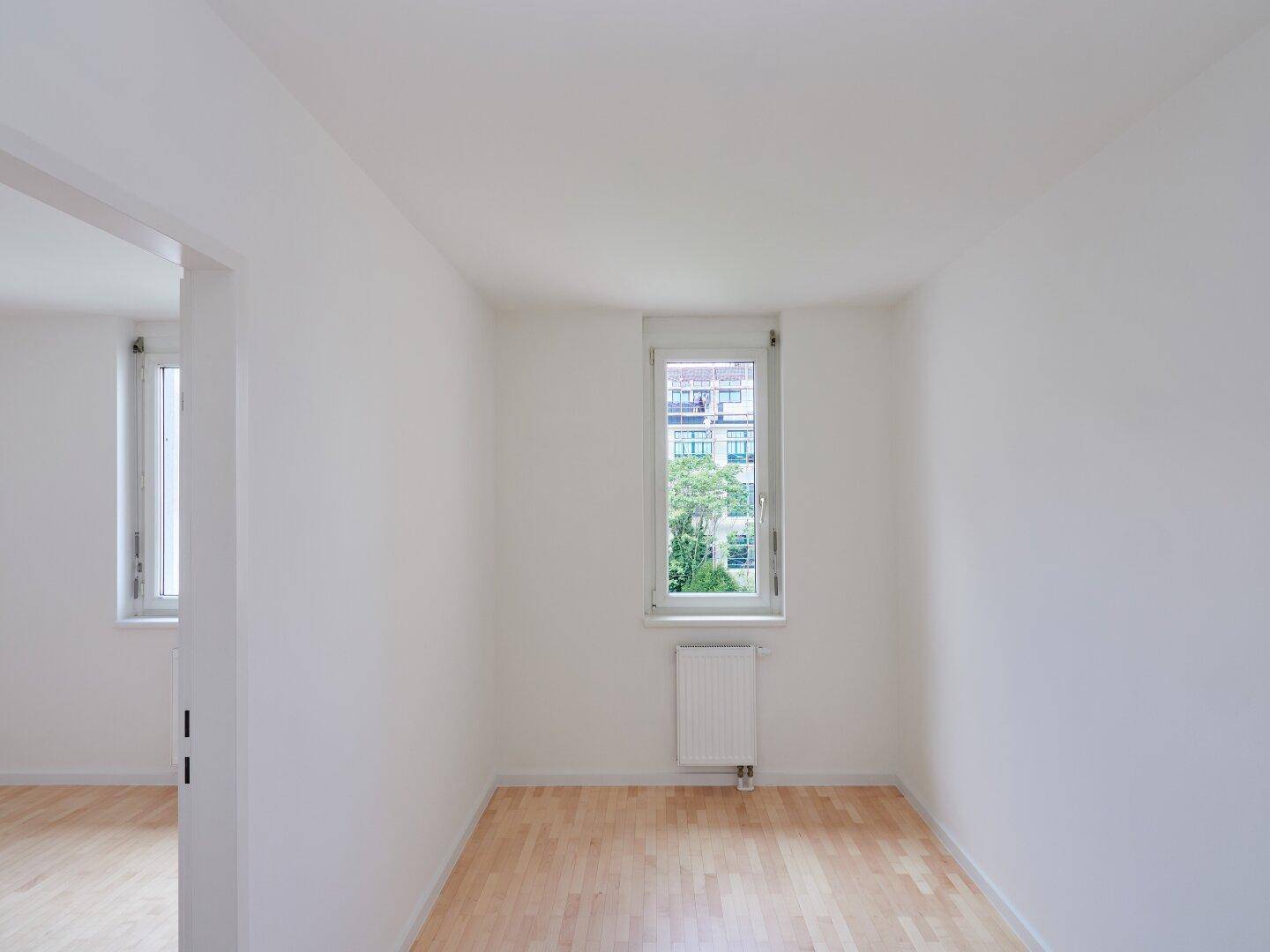 Helles Zimmer mit Holzboden, weißen Wänden und einem Fenster mit Blick ins Grüne.