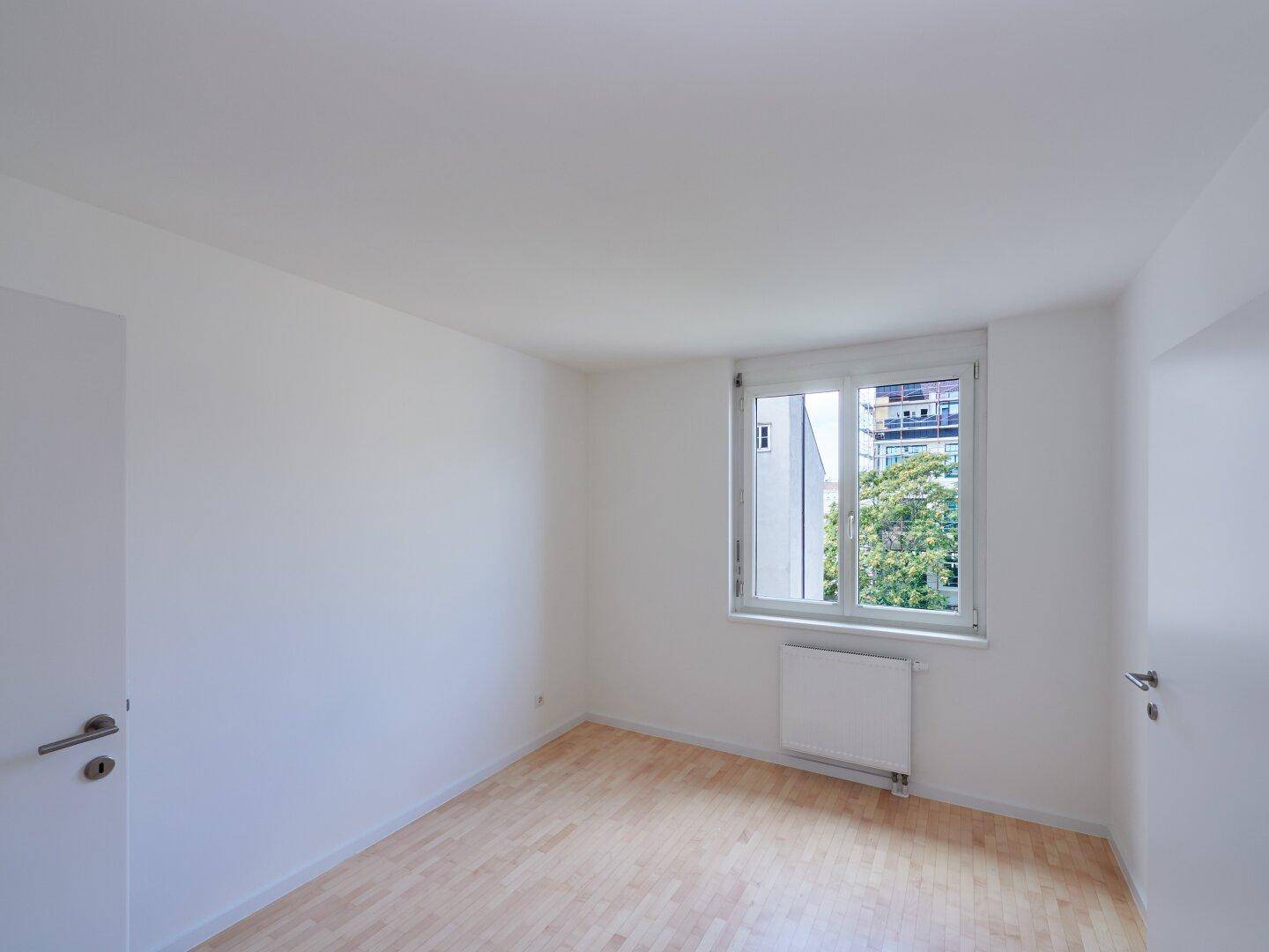Geräumiges Zimmer mit Holzboden, weißen Wänden und einem großen Fenster, das viel Licht hereinlässt.