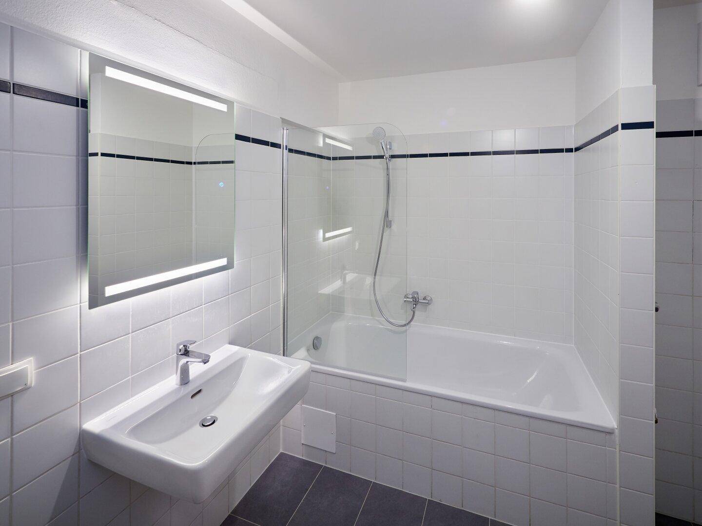 Modernes Badezimmer mit weißen Fliesen, Badewanne mit Duschwand und beleuchtetem Spiegel über dem Waschbecken.