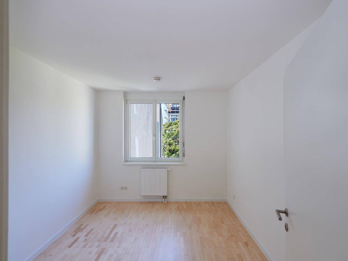 Helles Zimmer mit Holzboden, weißen Wänden und einem Fenster mit Blick ins Grüne.