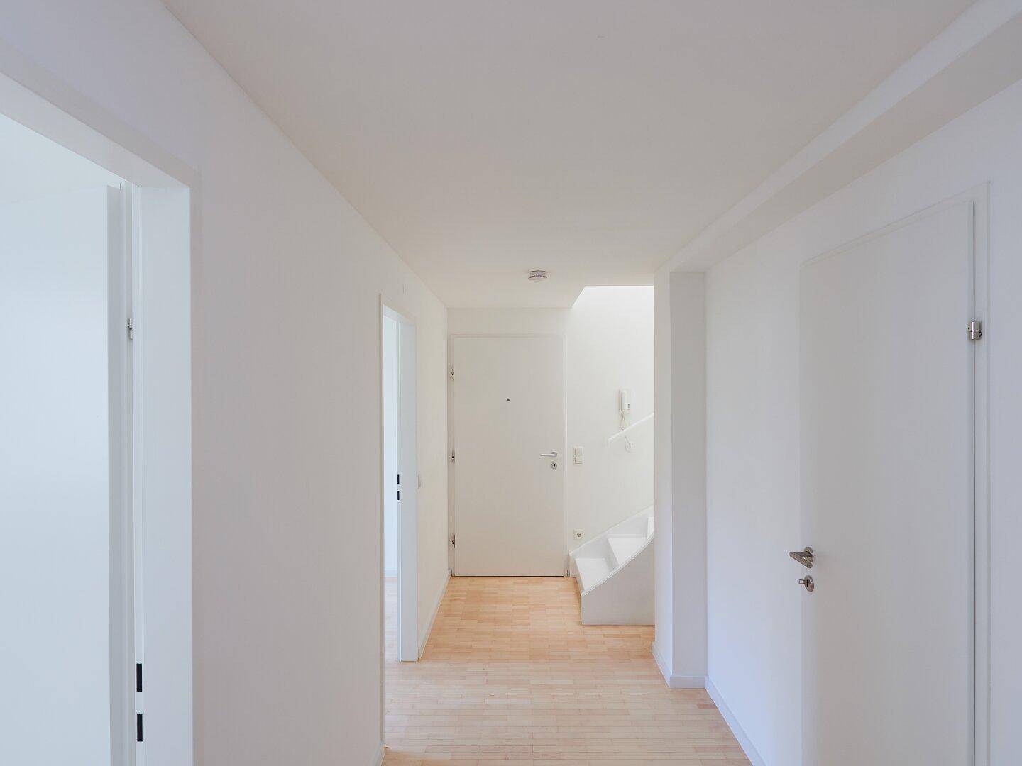 Heller Eingangsbereich mit Holzboden, weißen Wänden und Zugang zum Treppenhaus im Hintergrund.