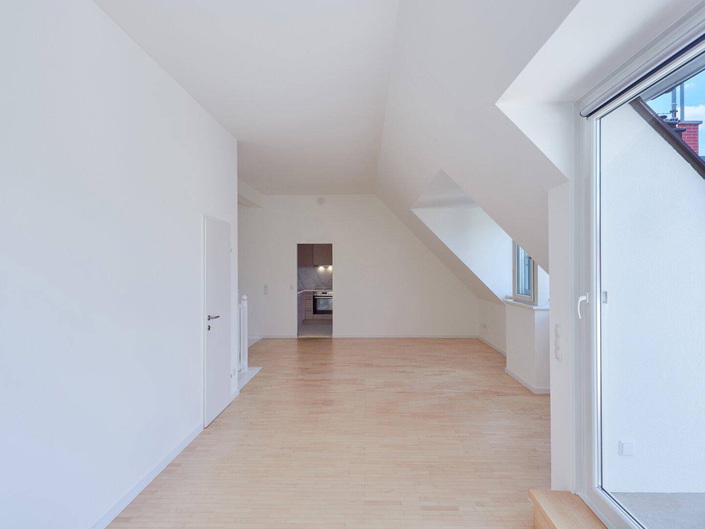 Offener Wohnbereich mit Holzboden, schrägen Wänden und Blick in die angrenzende Küche.