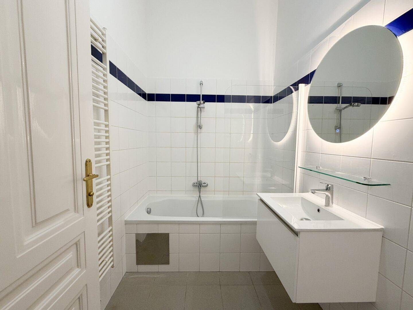Modernes Badezimmer mit Badewanne, Waschtisch und beleuchtetem Spiegel, akzentuiert durch eine blaue Fliesenlinie.