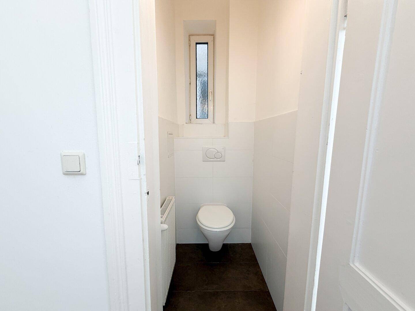 Separates WC mit wandhängender Toilette, kleinem Fenster und weißen Fliesen.