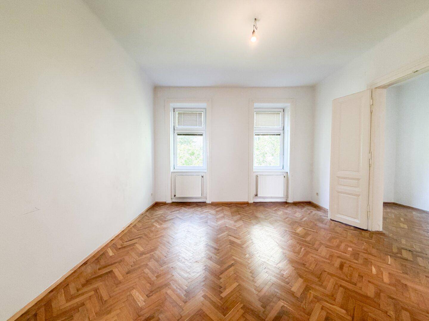 Helles Zimmer mit Fischgrätparkett, weißen Wänden und zwei Fenstern, ideal als Wohnraum.