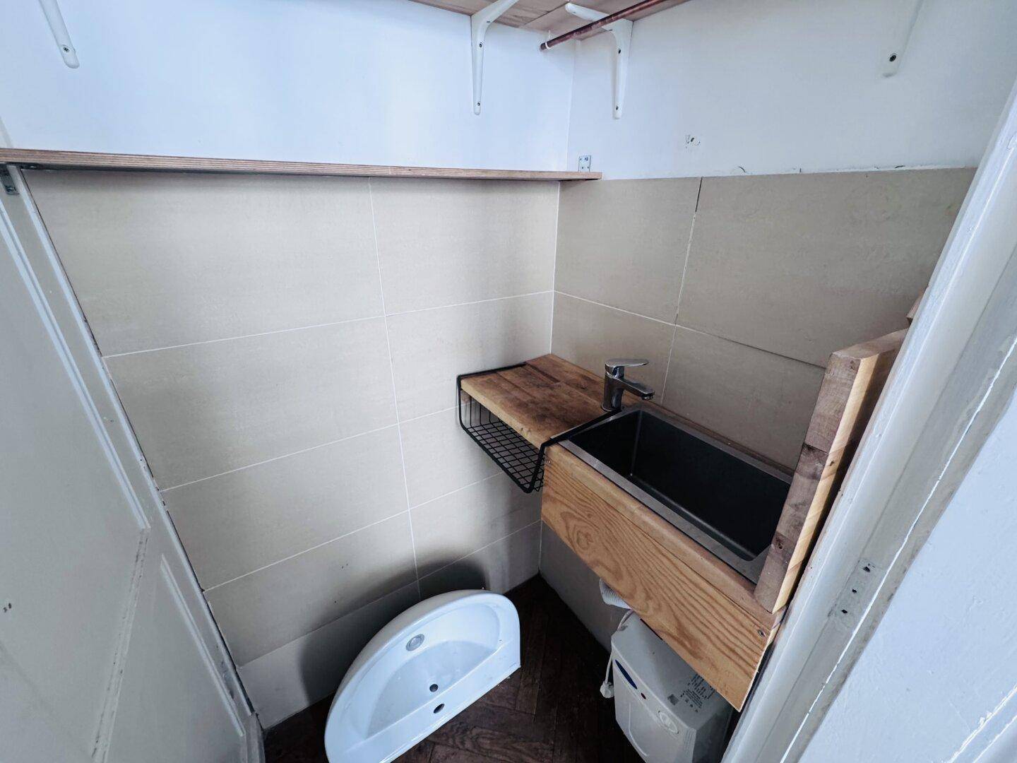 Kleiner, gefliester Toilettenraum mit einem Waschbecken auf einer Holzablage und einem weiteren Waschbecken am Boden.