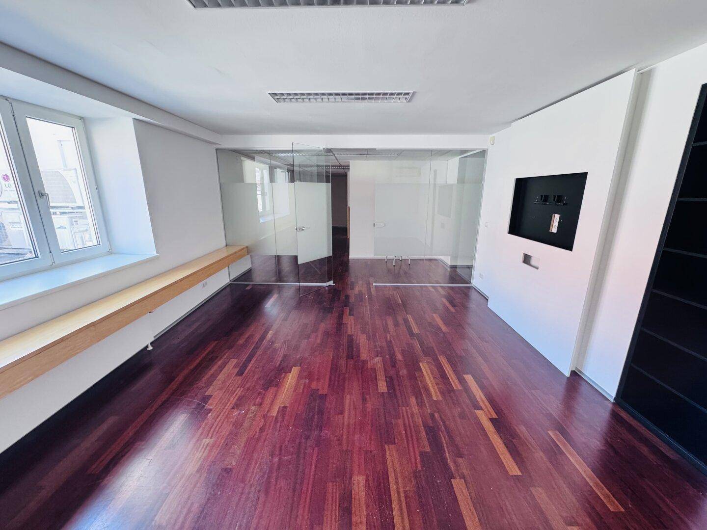 Helles Büro mit dunklem Holzparkett, weißen Wänden und einer modernen Wandhalterung für einen Bildschirm.