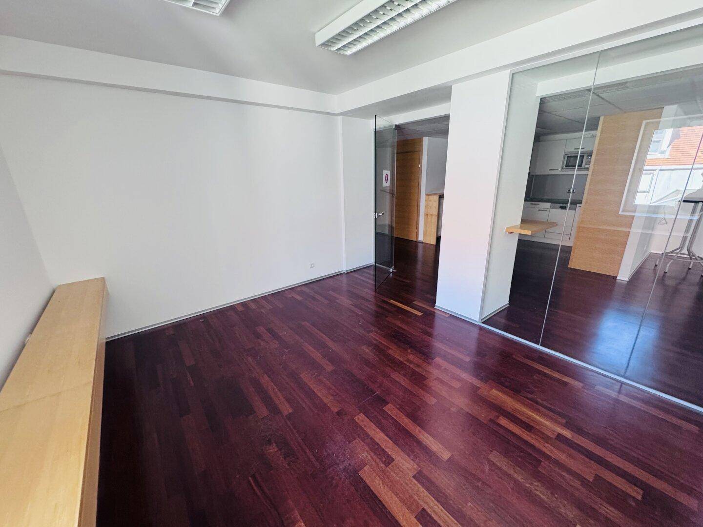 Leeres Büro mit dunklem Parkettboden, weißen Wänden und einer langen Holzablage unter dem Fenster.