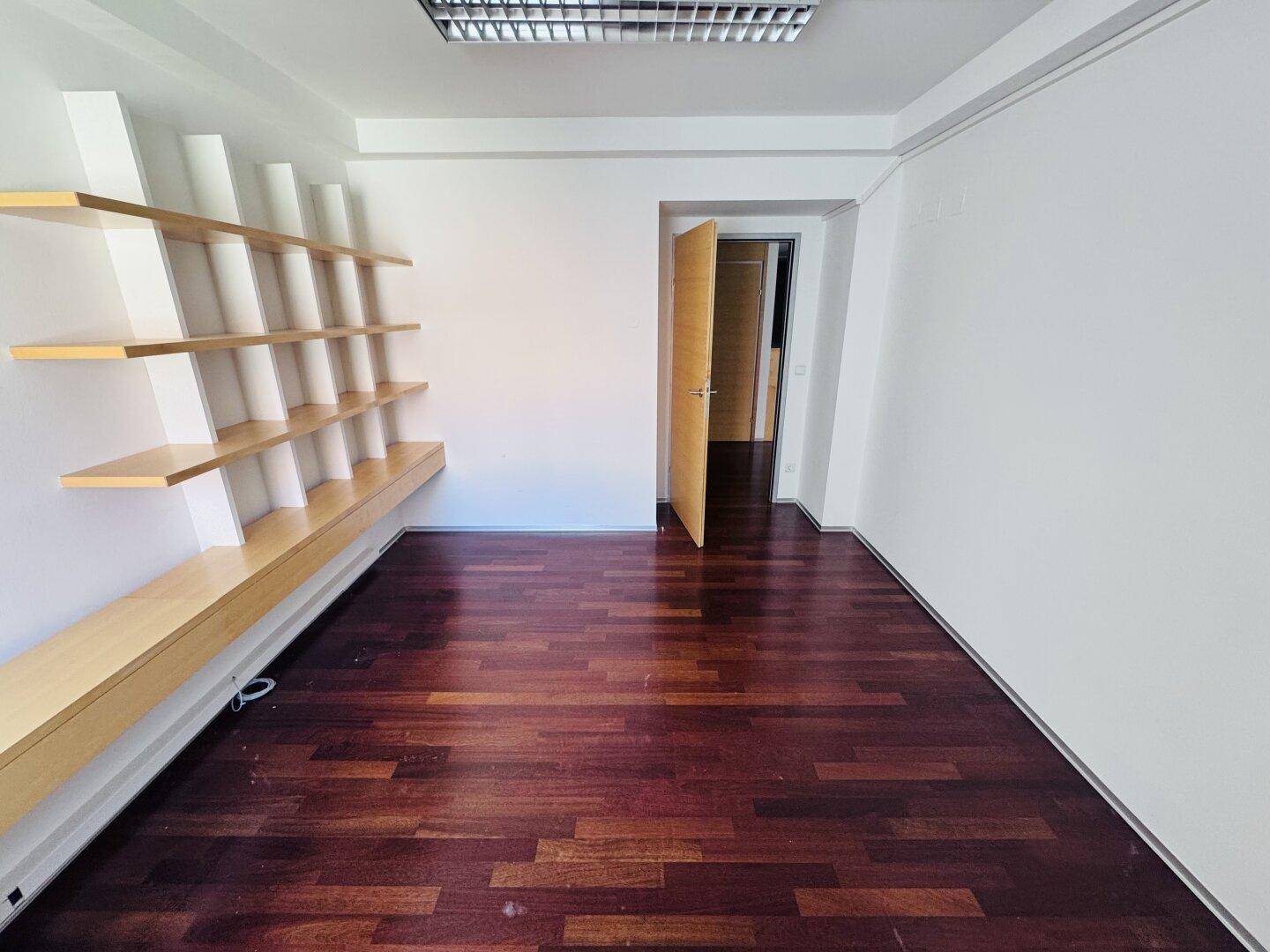 Helles Büro mit dunklem Parkettboden, weißen Wänden und einem modernen, eingebauten Holzregalsystem.