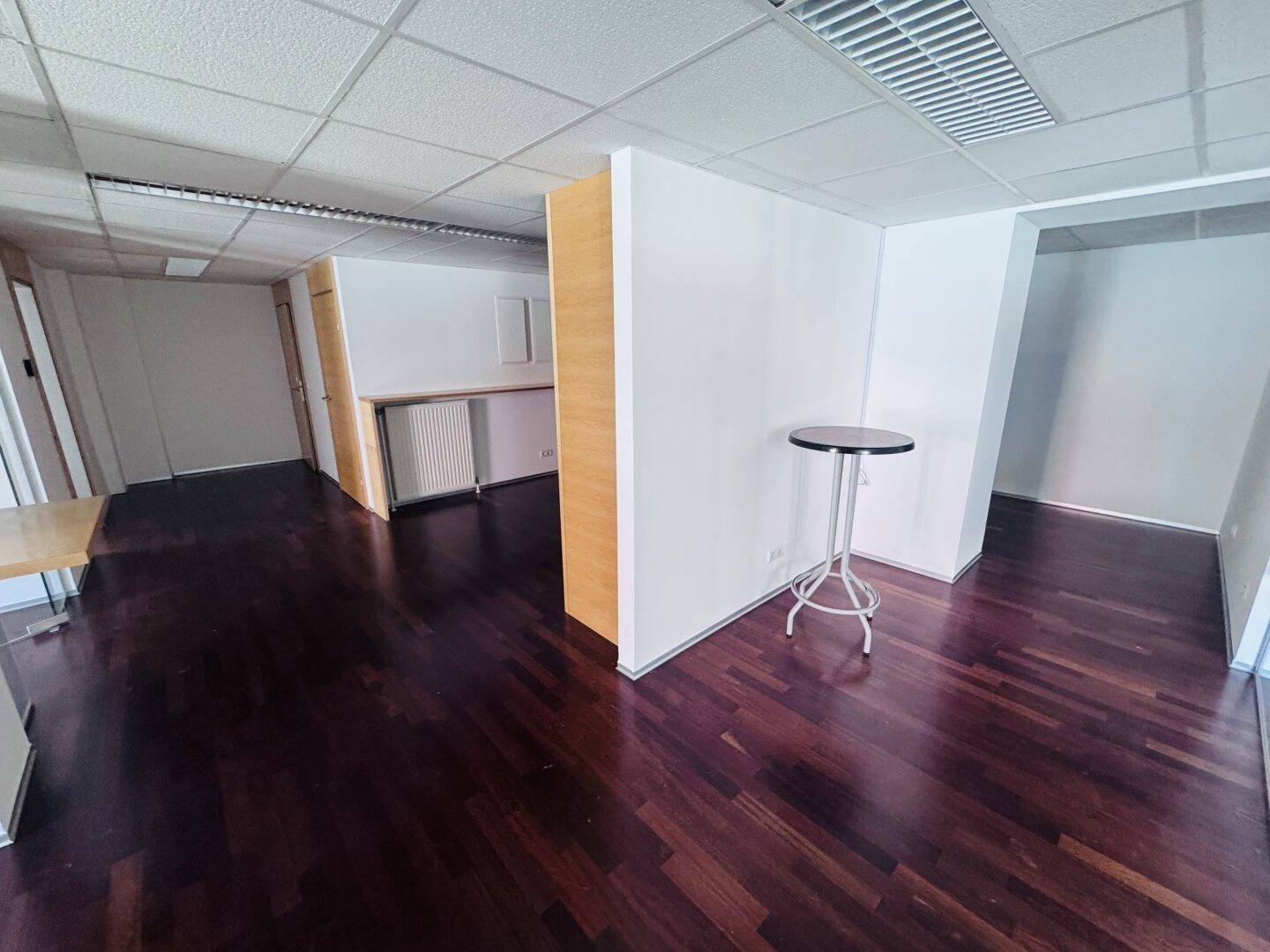Offener Bürobereich mit dunklem Parkettboden, weißen Wänden und einem kleinen Stehtisch, der den Raum belebt.