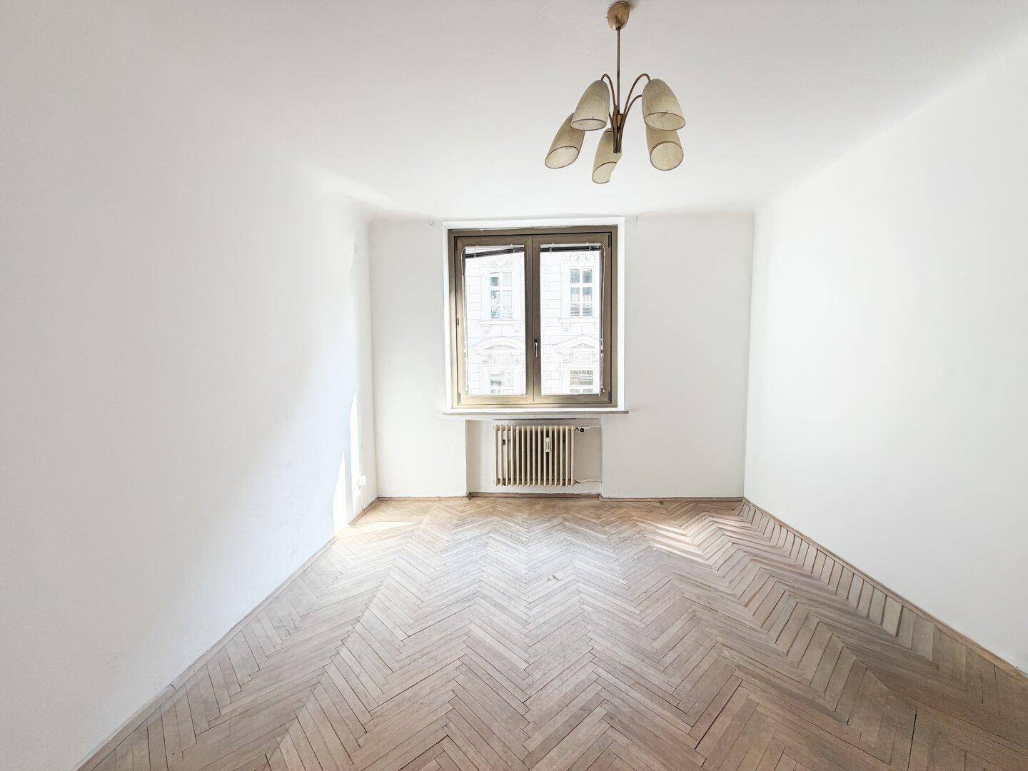 Lichtdurchflutetes Zimmer mit Fischgrätparkett, einem Fenster und einem Heizkörper, bietet viel Tageslicht.