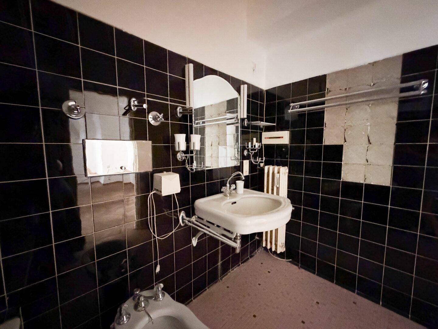 Badezimmer mit schwarzen Wandfliesen, Waschbecken, Spiegel und Heizkörper.