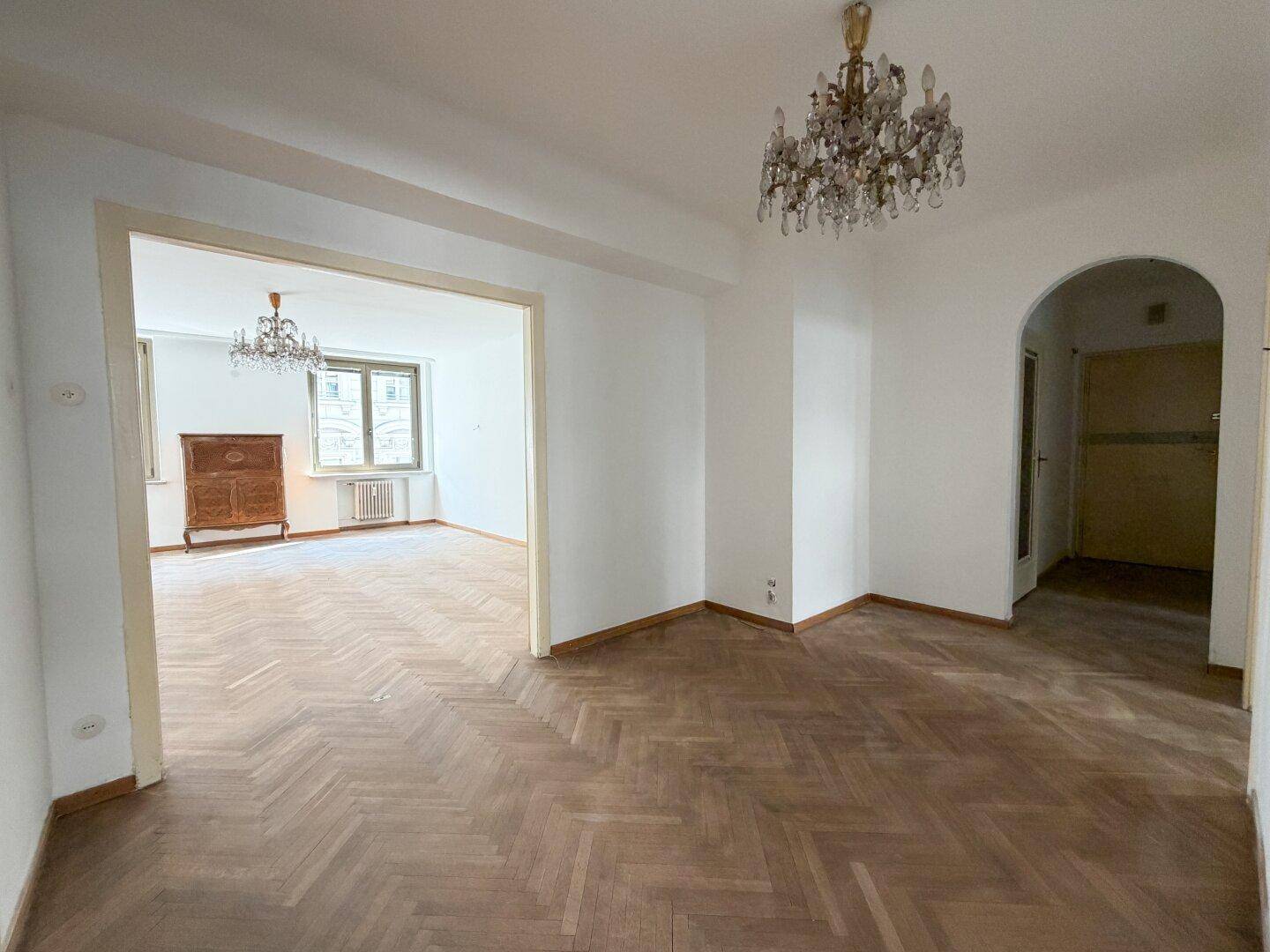 Geräumiges Wohnzimmer mit Fischgrätparkett, hohen Decken und elegantem Kronleuchter, bereit zur individuellen Gestaltung.