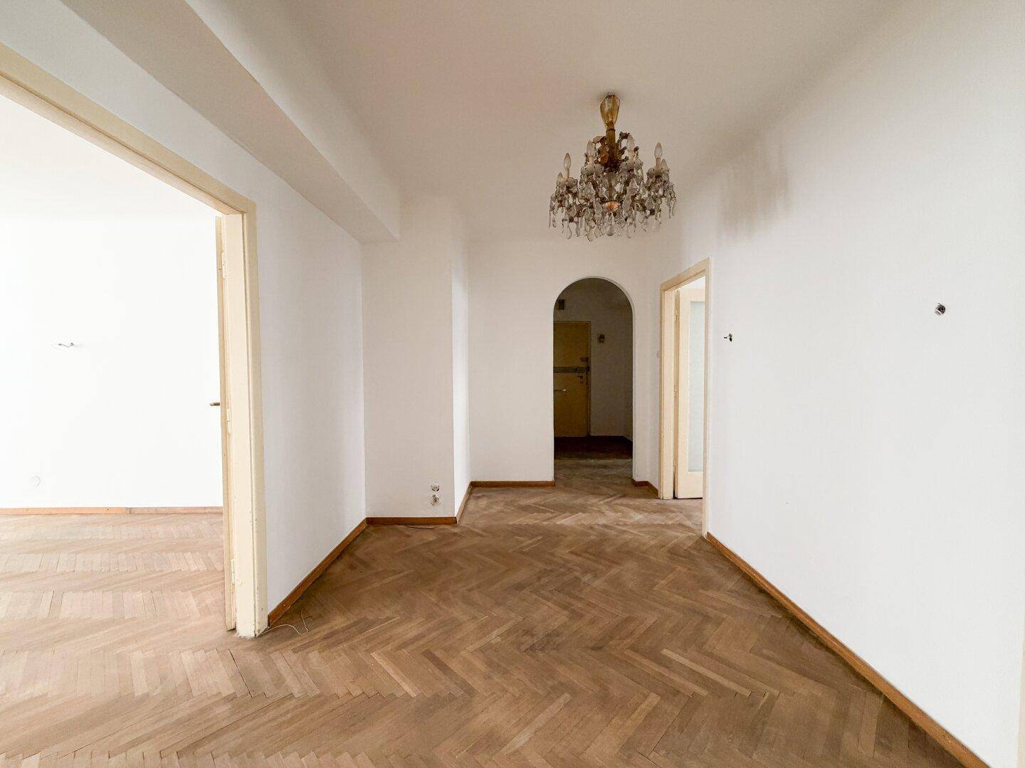 Helles Zimmer mit Fischgrätparkett, Kronleuchter und einem eleganten Rundbogen zu einem weiteren Bereich.