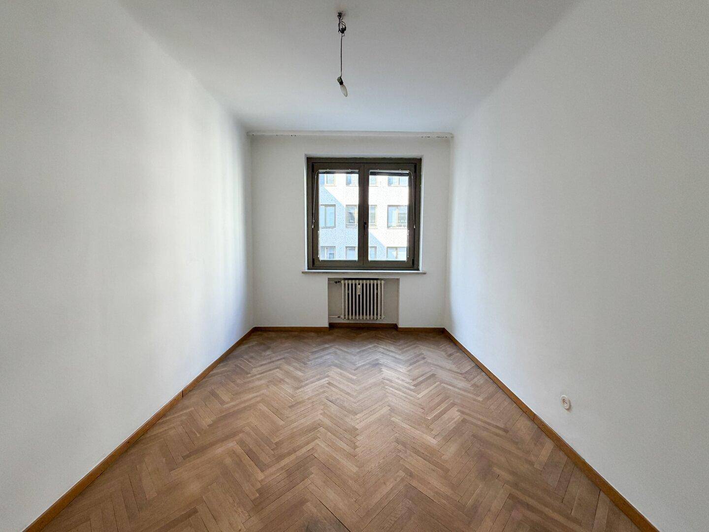 Kleineres Zimmer mit Fischgrätparkett, einem Fenster und weiß gestrichenen Wänden.