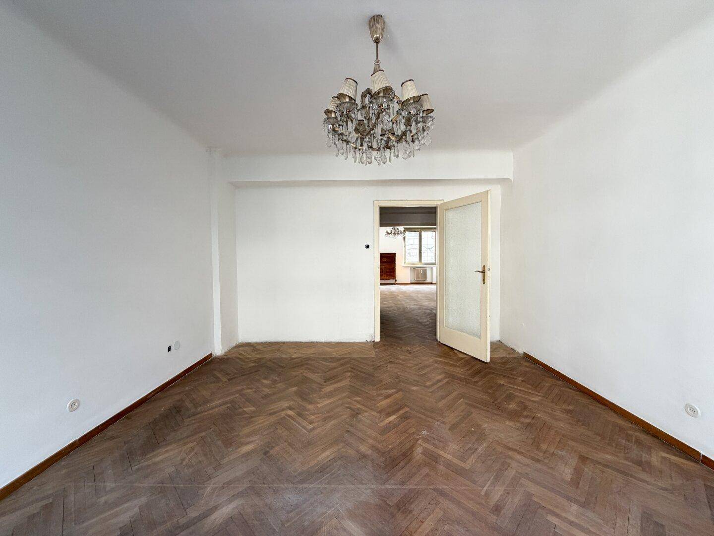 Heller Raum mit Fischgrätparkett und offenem Durchgang zu einem weiteren Zimmer.