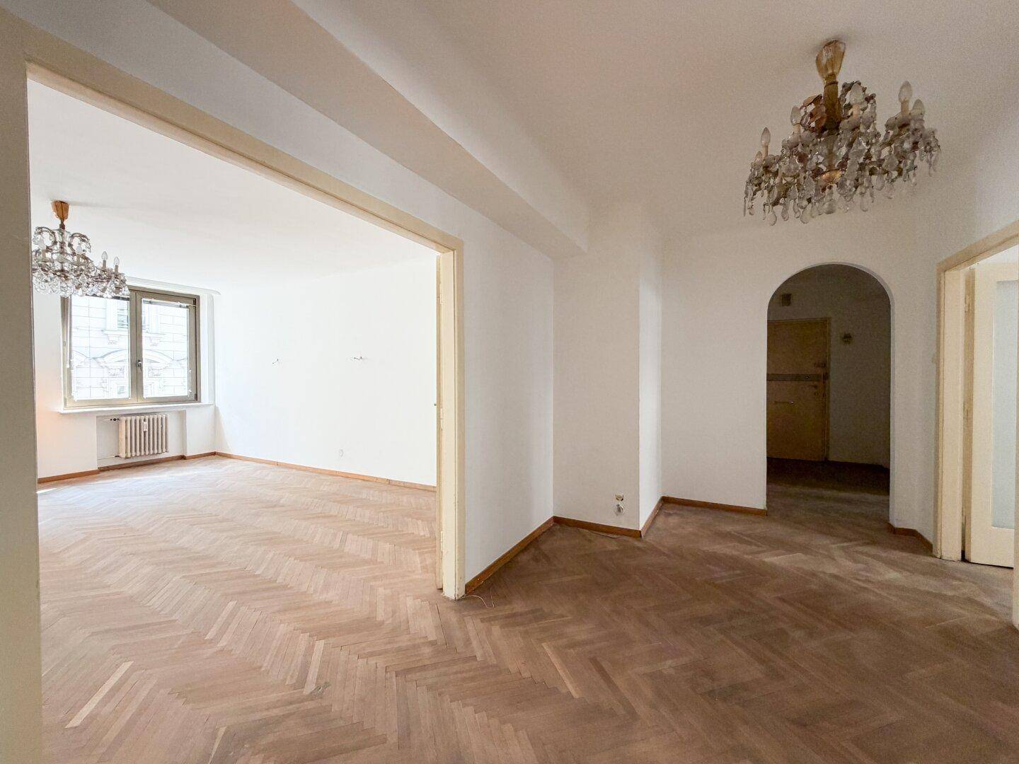 Großzügiger Wohnbereich mit Fischgrätparkett und offenem Übergang zu einem weiteren Zimmer.