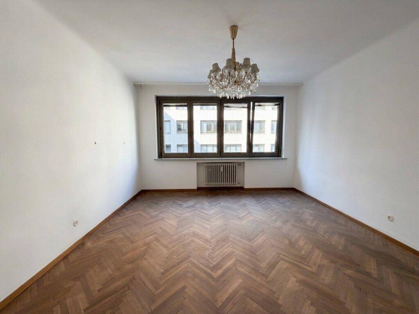 Geräumiges Zimmer mit Fischgrätparkett, großem Fenster und einem klassischen Kronleuchter.