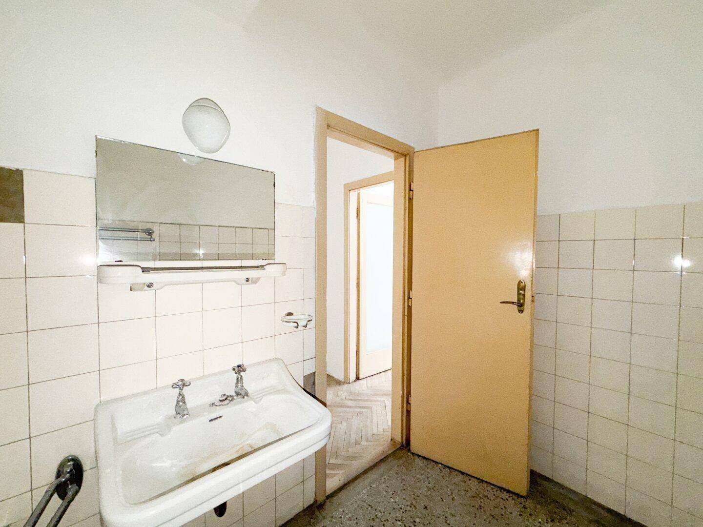 Badezimmer mit Waschbecken, Spiegel und weißen Fliesen, das durch eine offene Tür zugänglich ist.